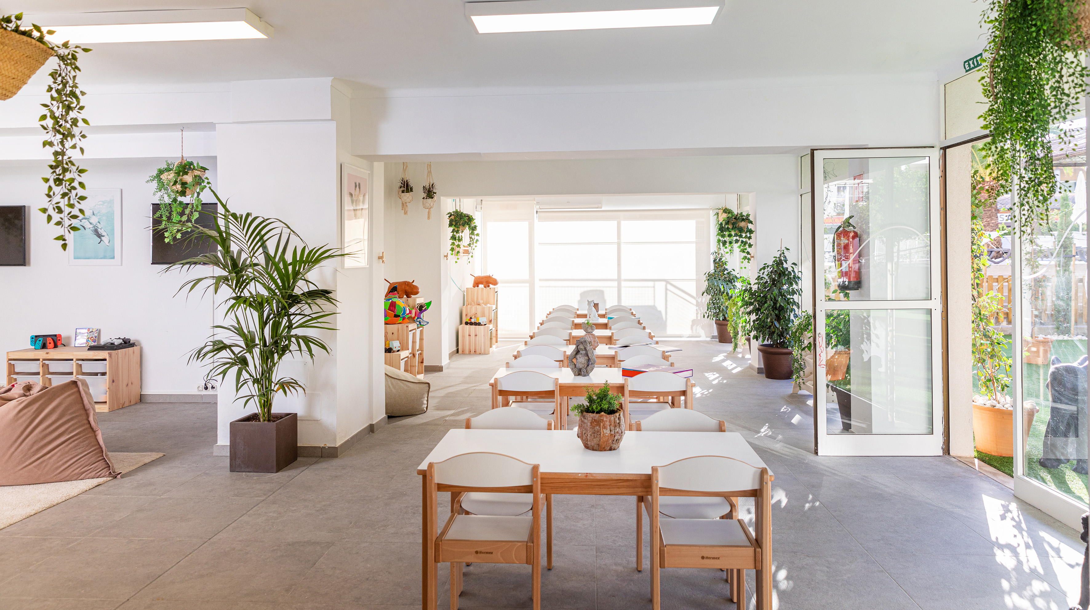 Bright children's classroom with small tables, chairs, play area, and lush green plants.