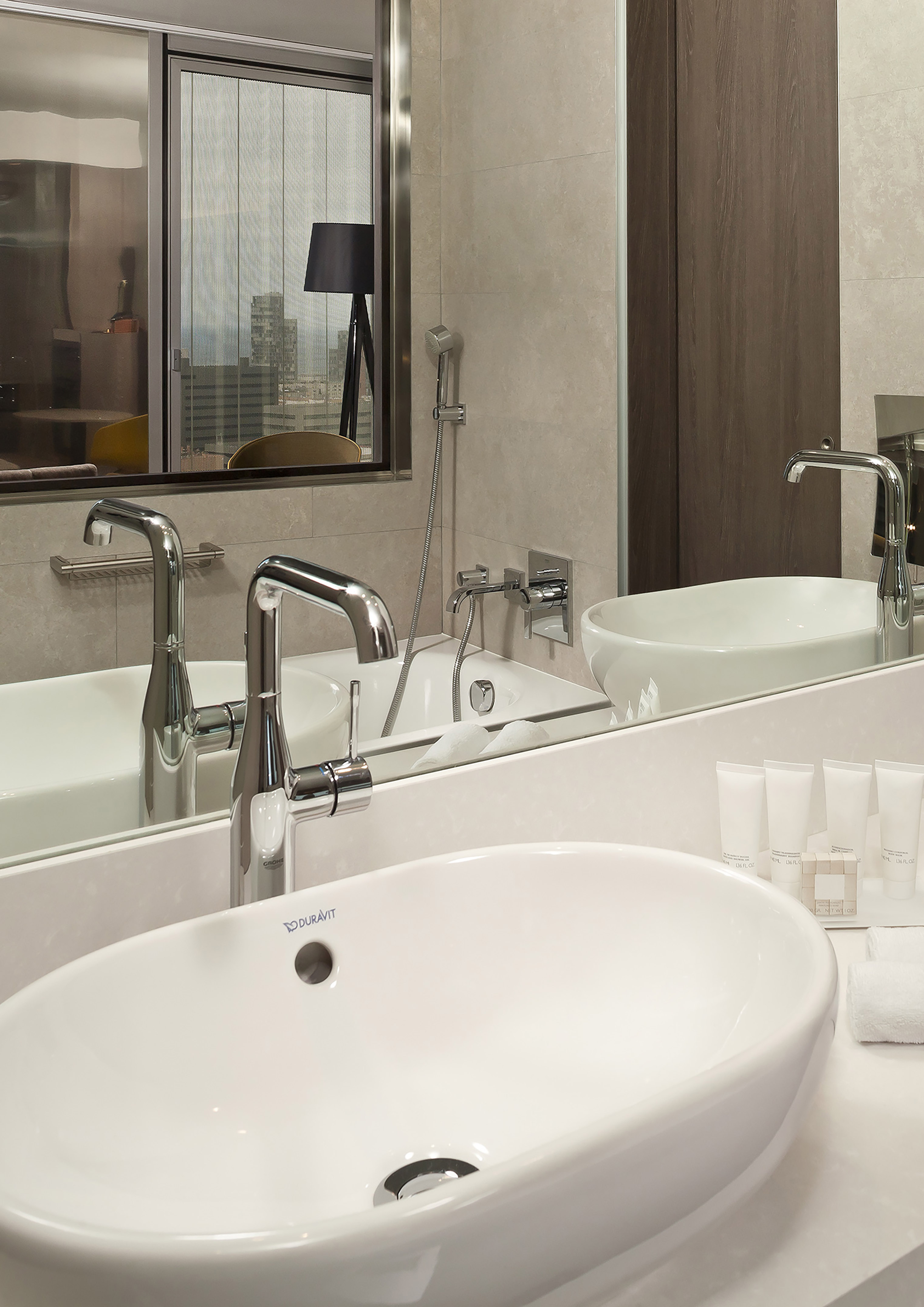 Modern hotel bathroom vanity with white sink, chrome faucet, city view.