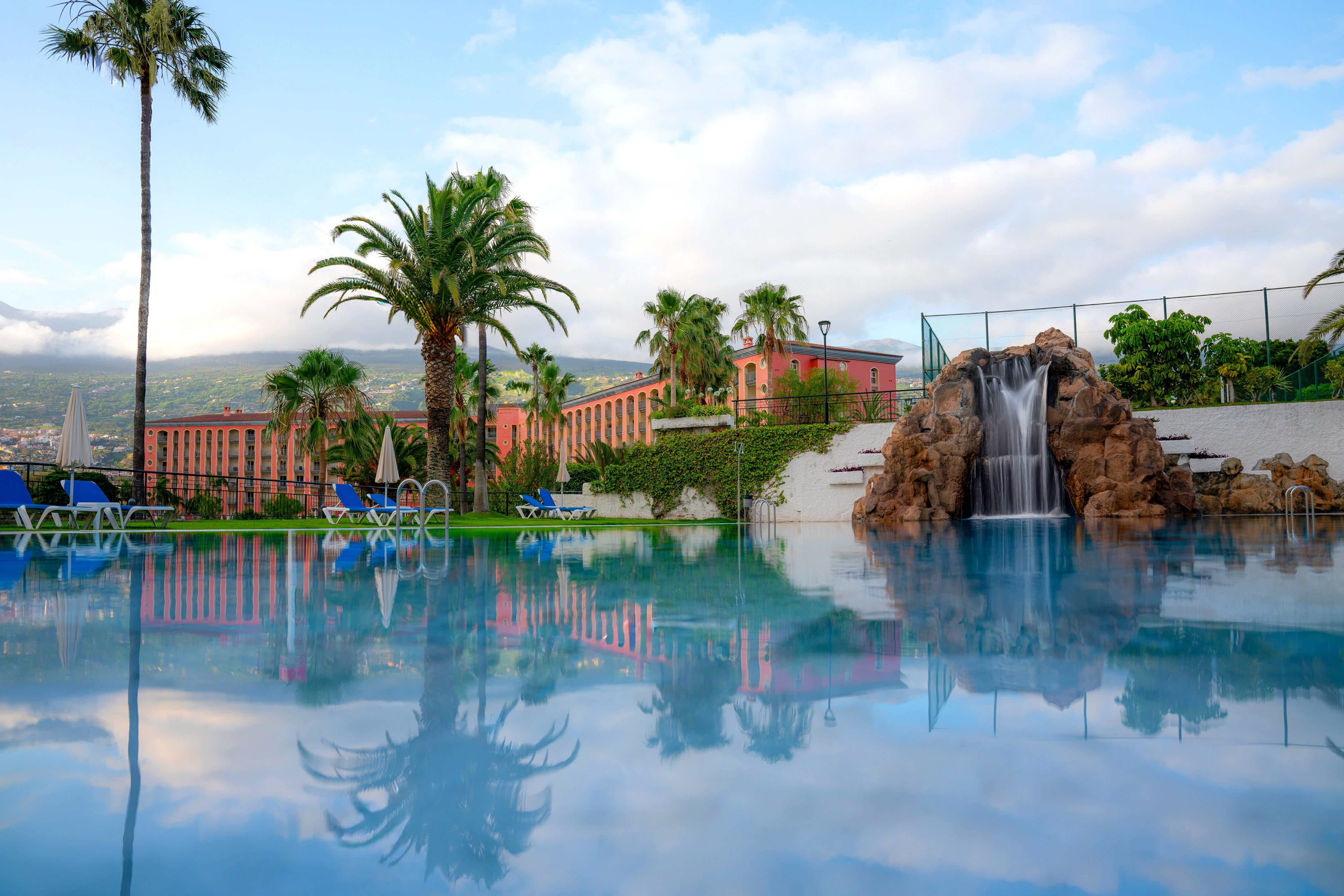 Tropical hotel pool with rock waterfall, palm trees, and pink building.