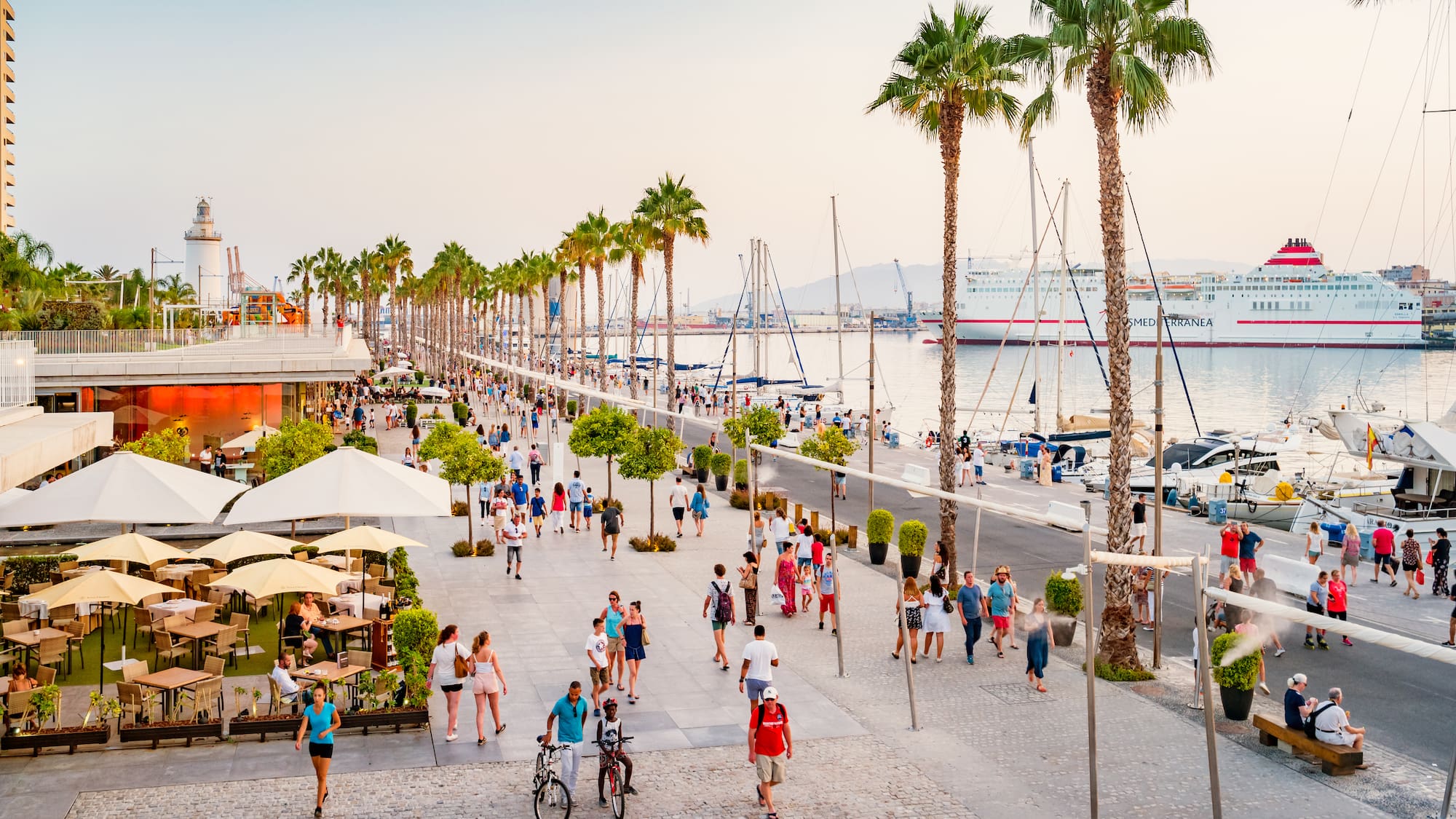 a group of people walking on a sidewalk with palm trees and boats