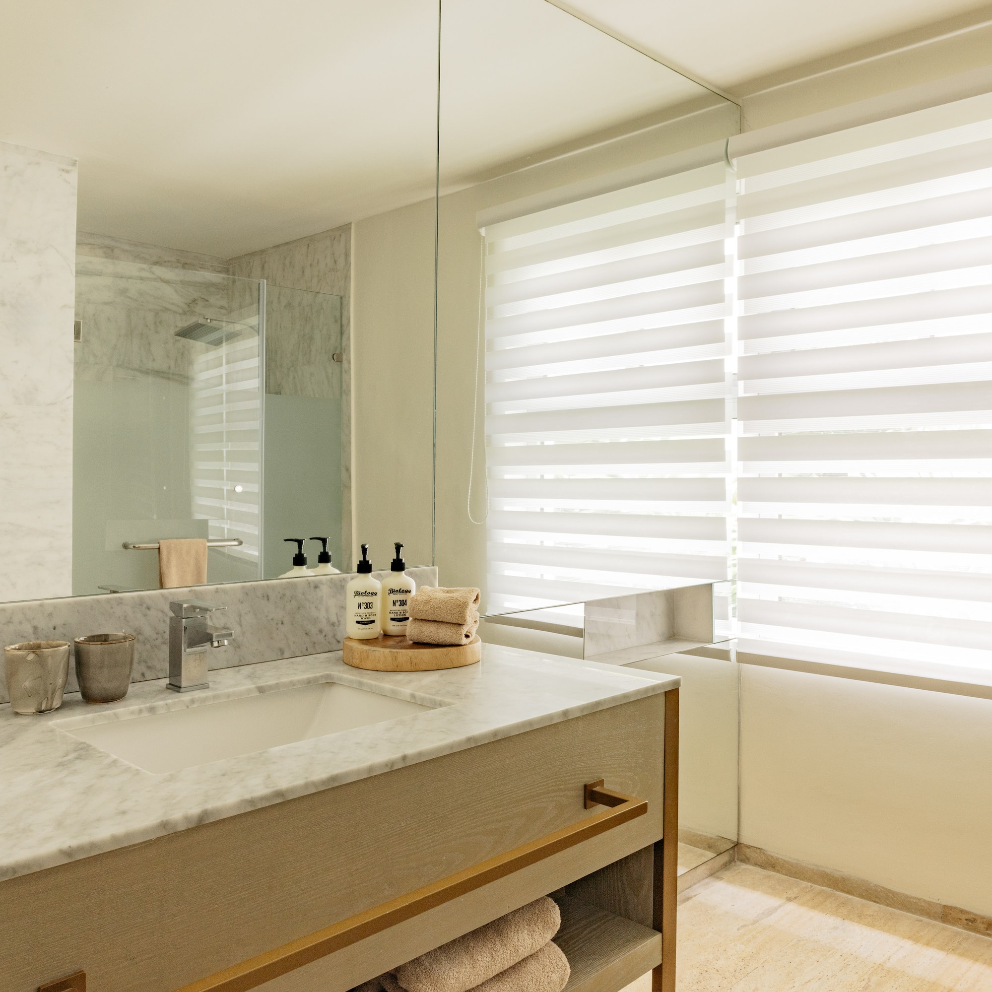 Modern bathroom with marble vanity, large mirror, glass shower, and window blinds.