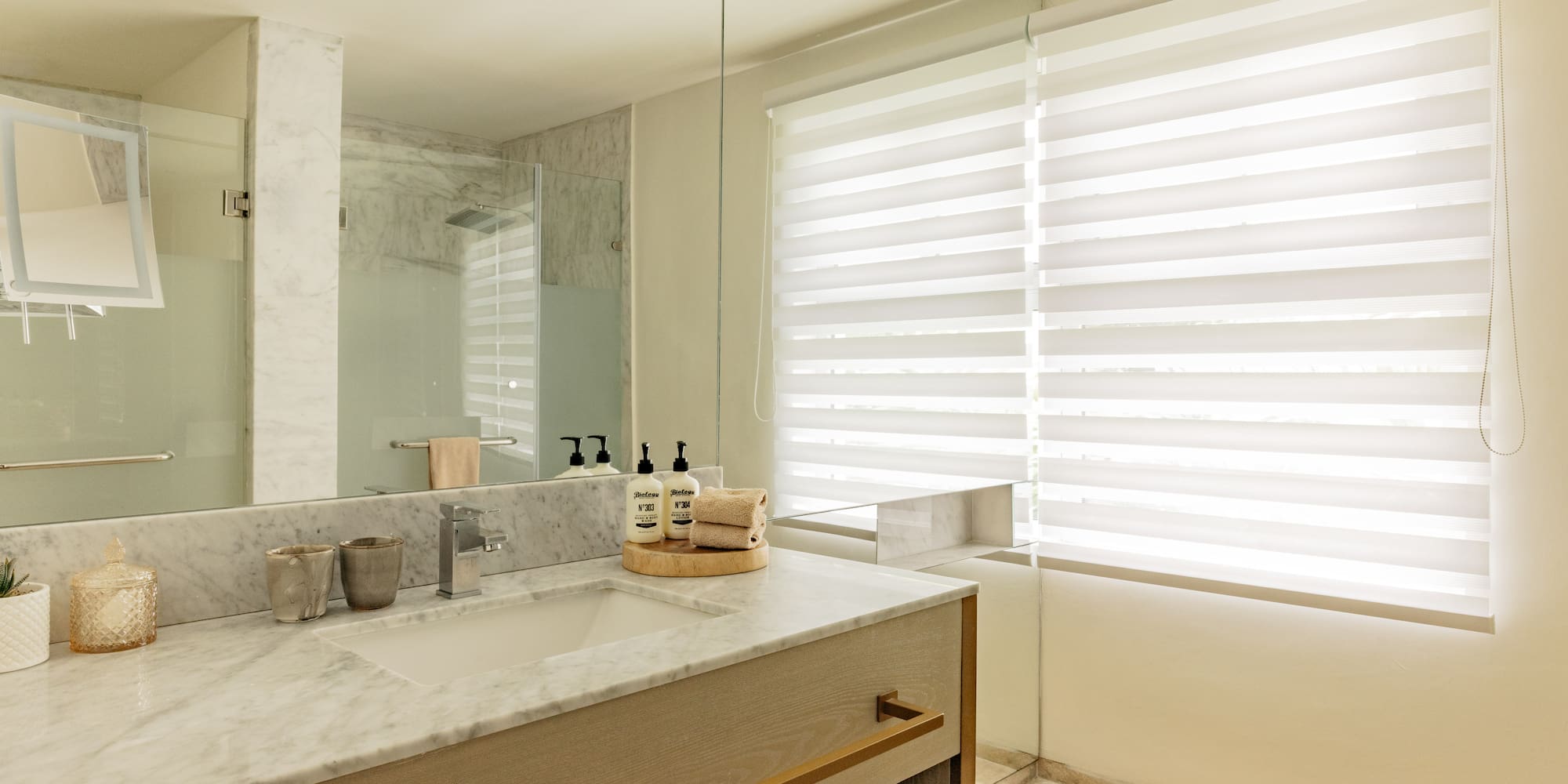 Modern bathroom with marble vanity, large mirror, glass shower, and window blinds.