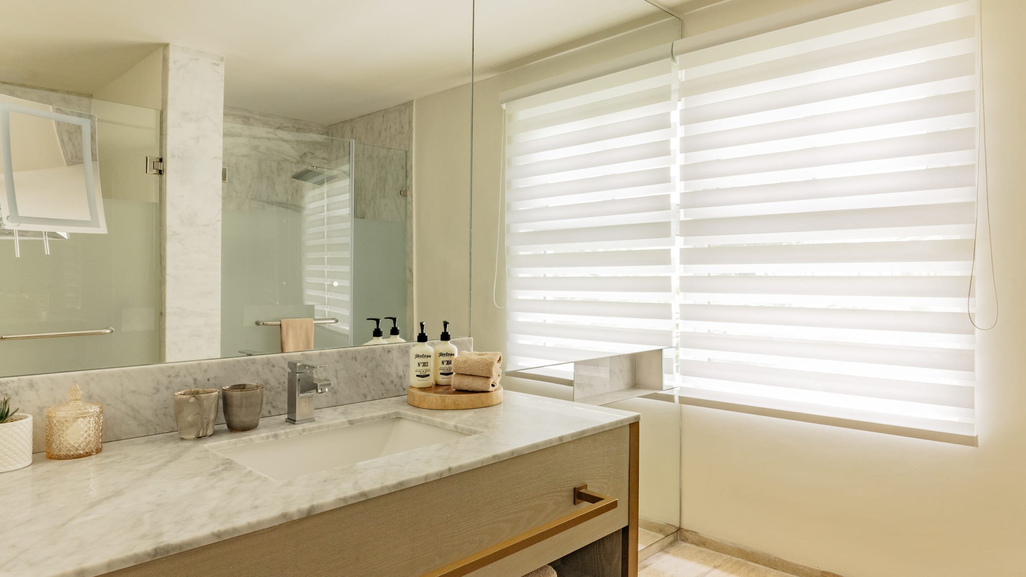 Modern bathroom with marble vanity, large mirror, glass shower, and window blinds.