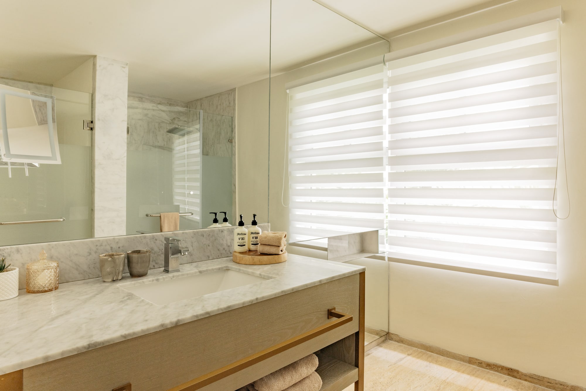 Modern bathroom with marble vanity, large mirror, glass shower, and window blinds.