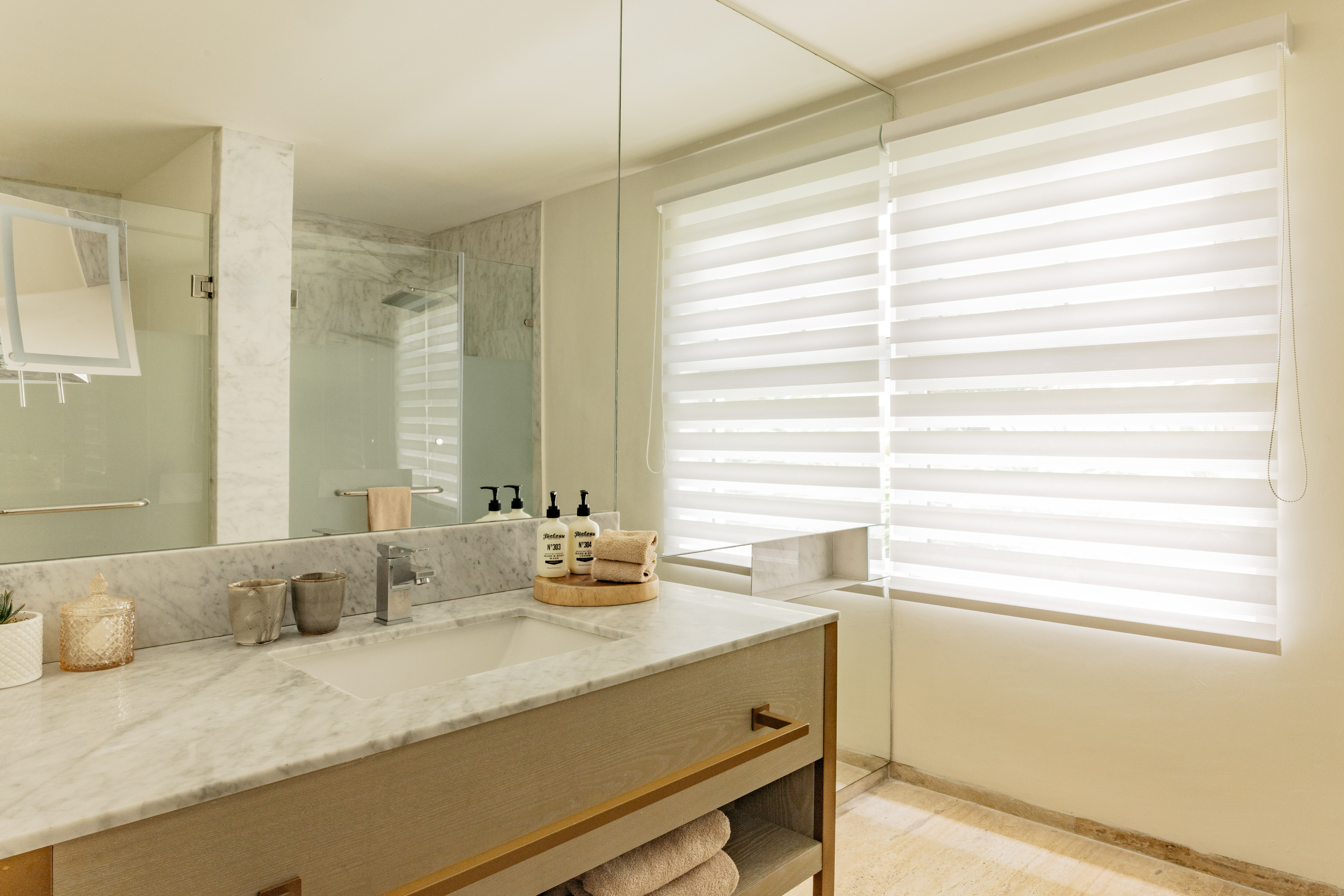 Modern bathroom with marble vanity, large mirror, glass shower, and window blinds.