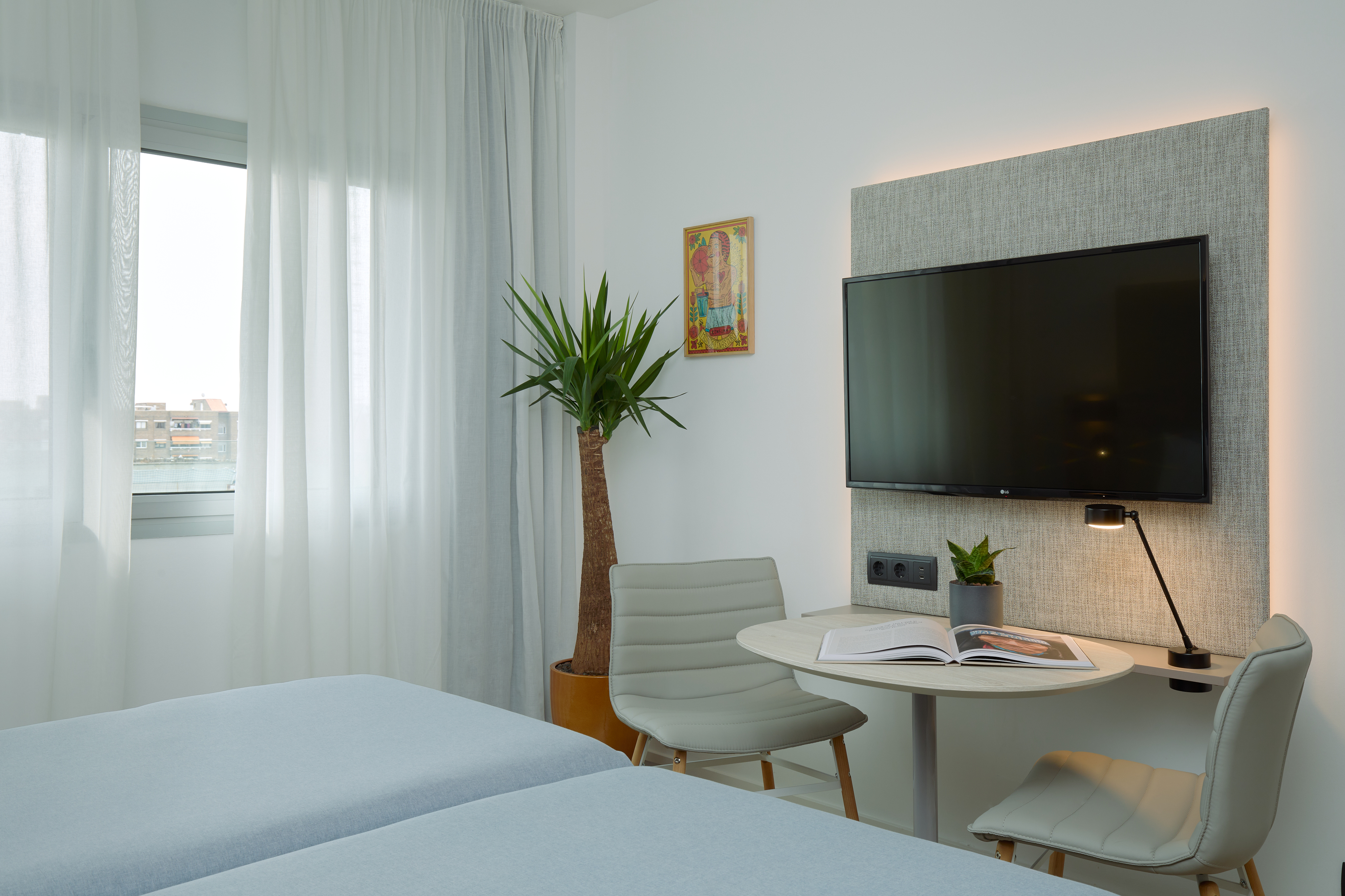 Bright modern hotel room with twin beds, wall TV, desk, and sheer window.