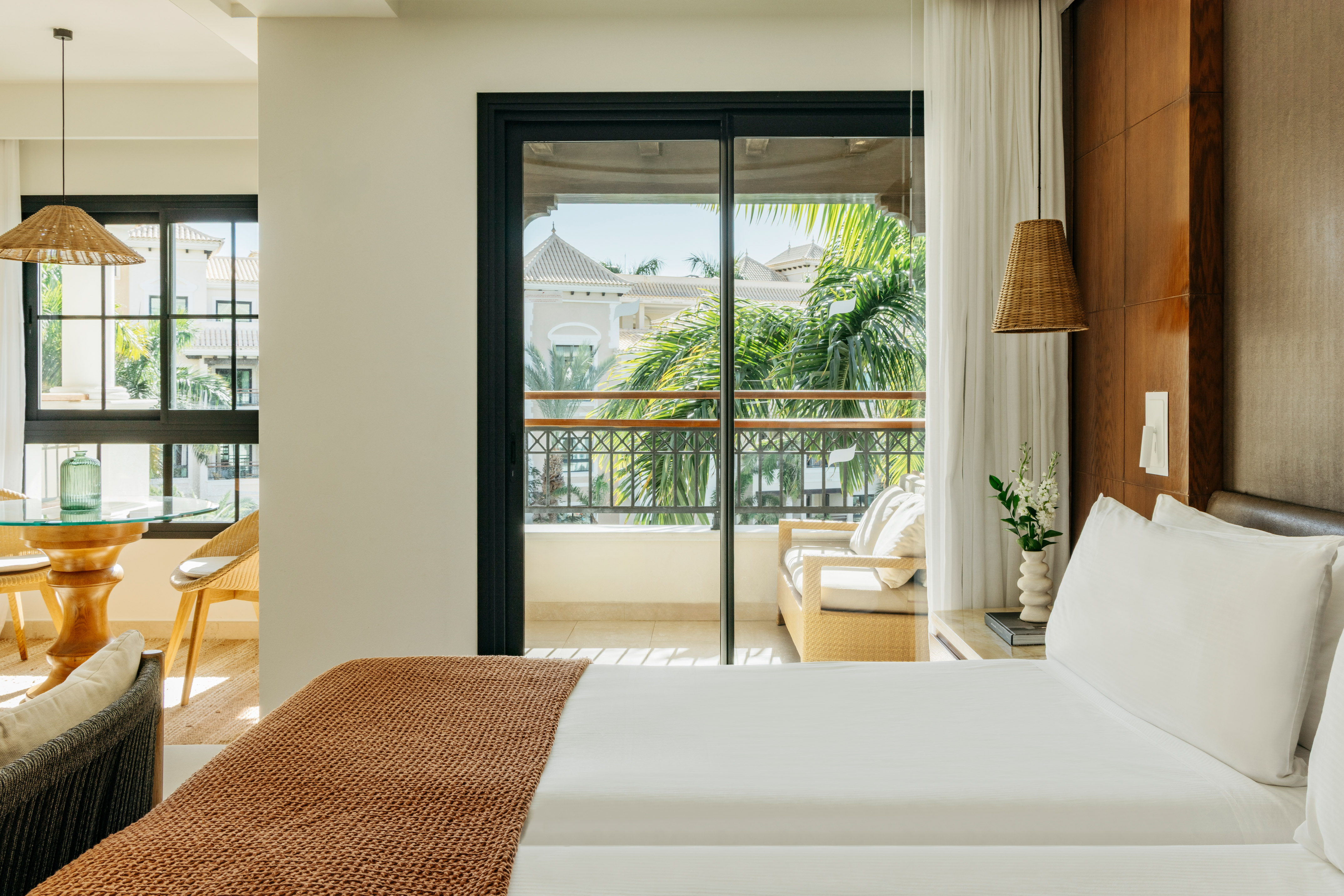 Bright hotel room with bed, balcony overlooking resort, and lush palm trees.