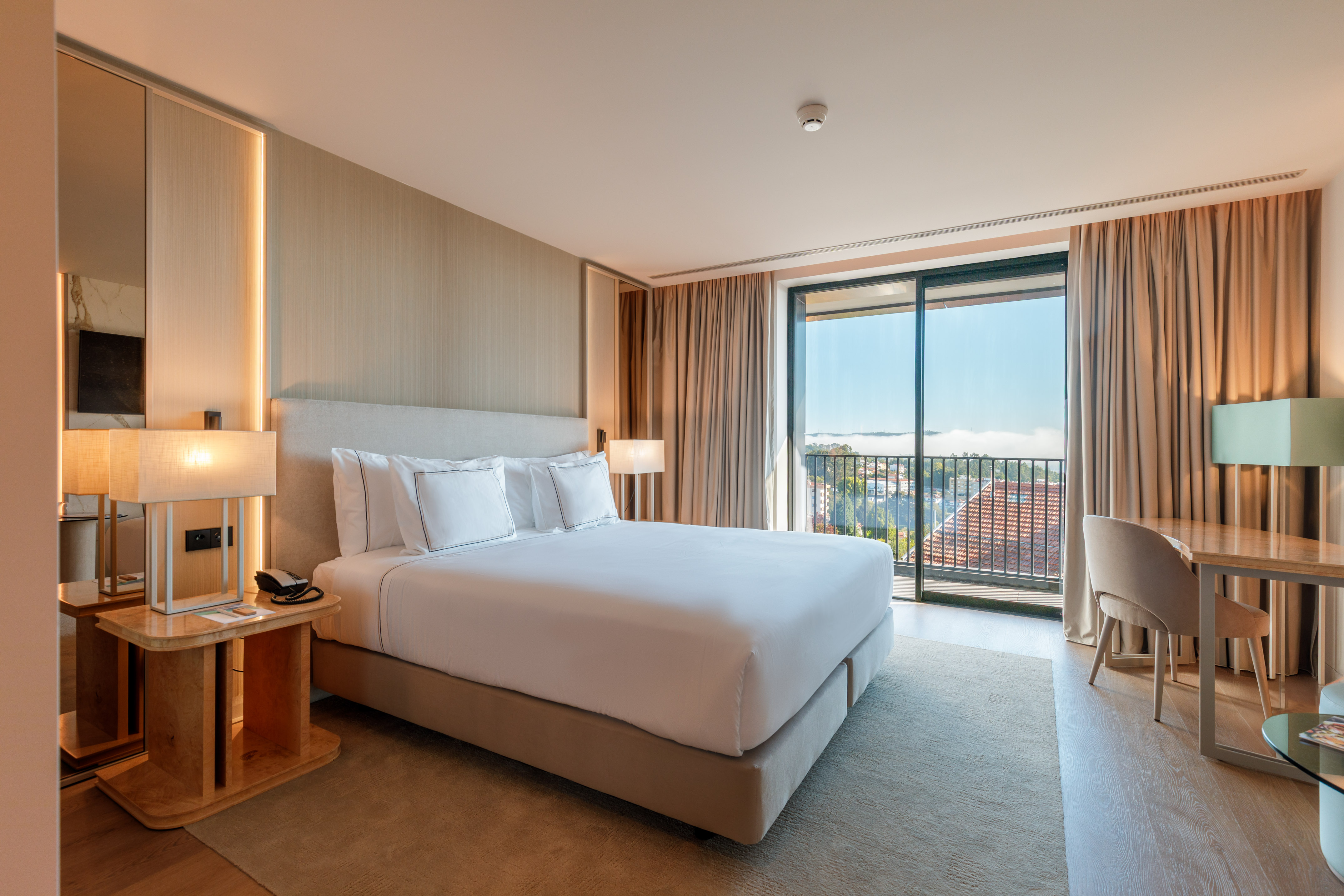 Modern hotel bedroom with large bed, desk, and balcony offering city view.