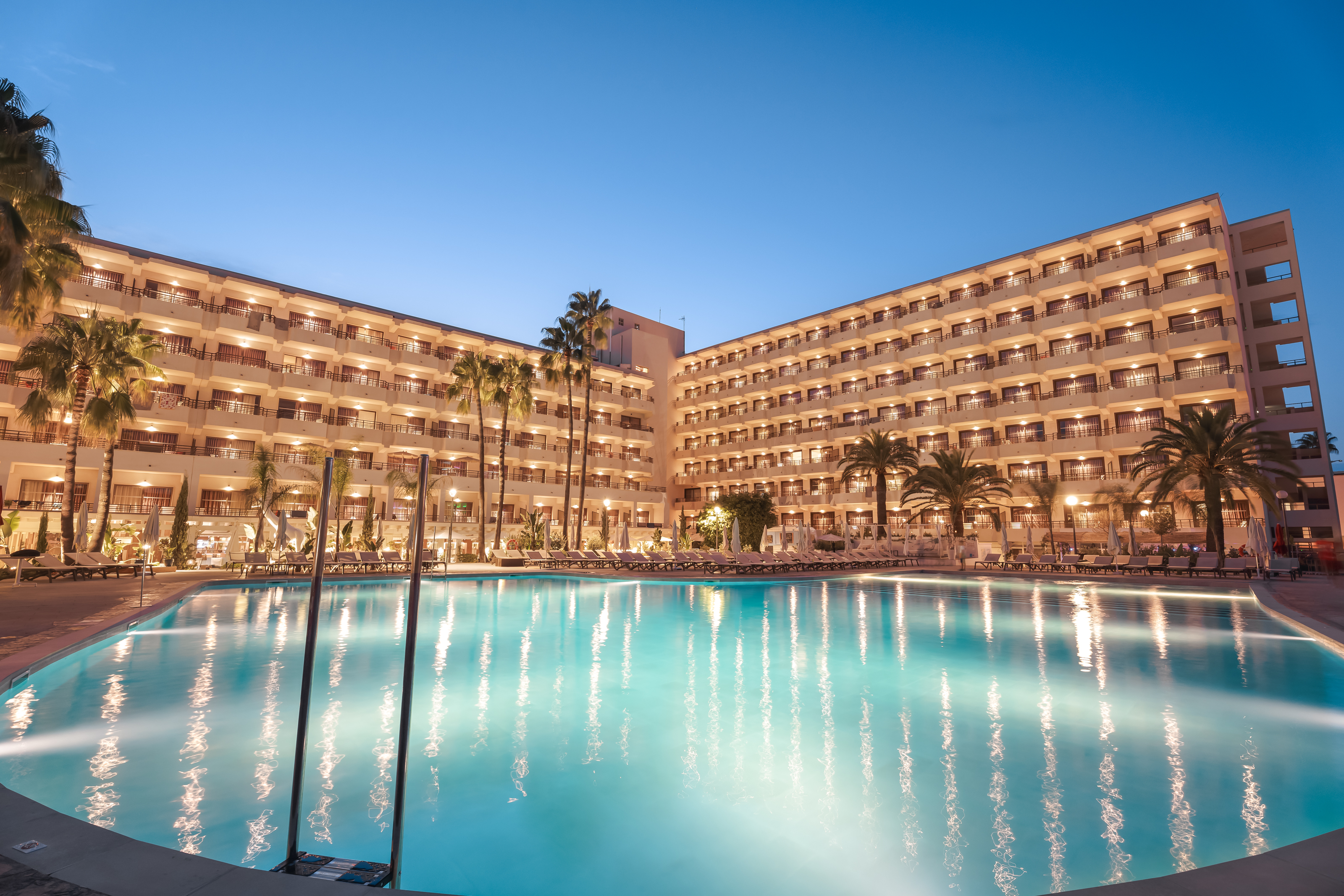 Illuminated resort hotel with large swimming pool and palm trees at evening.