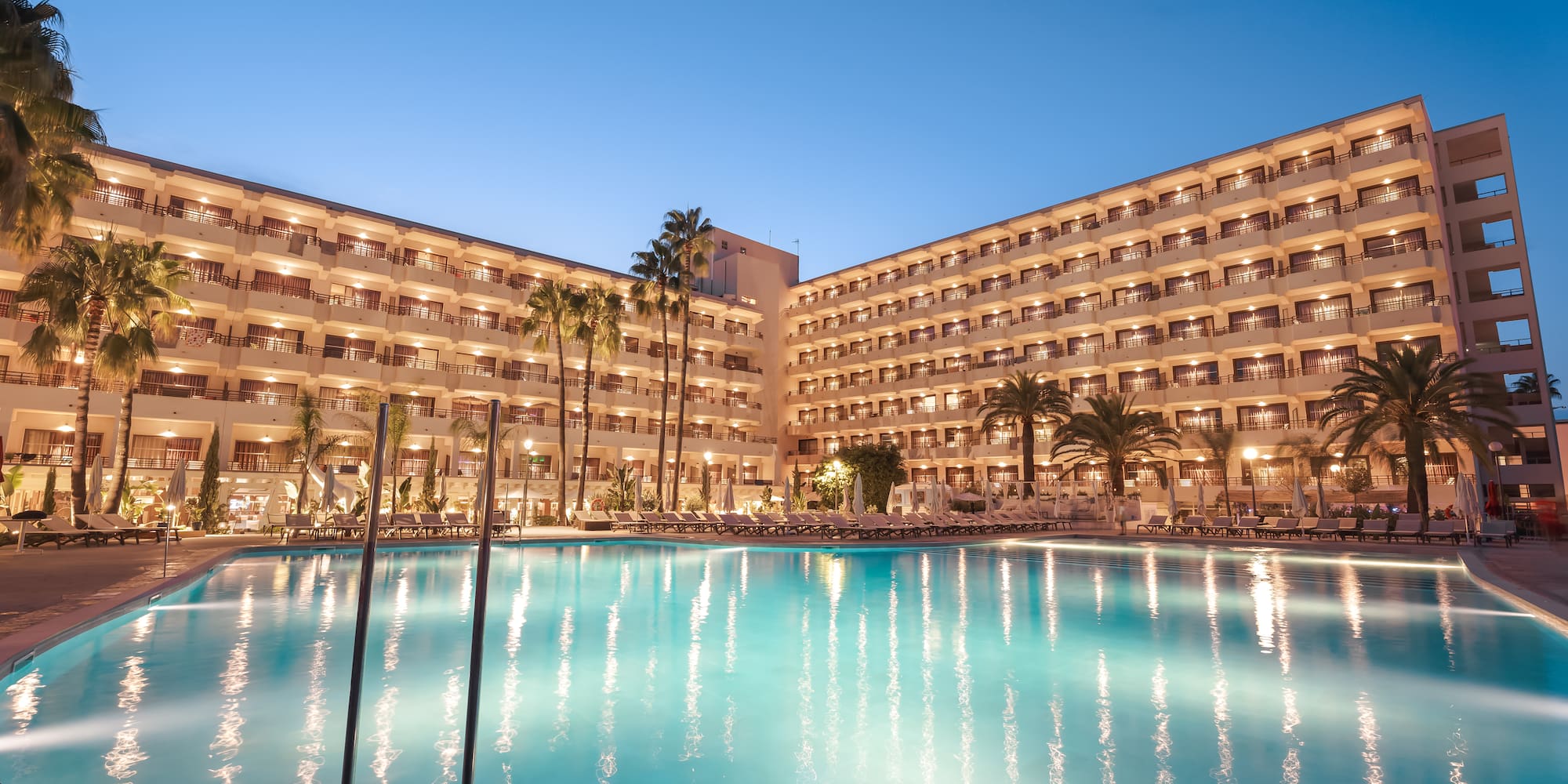 Illuminated resort hotel with large swimming pool and palm trees at evening.