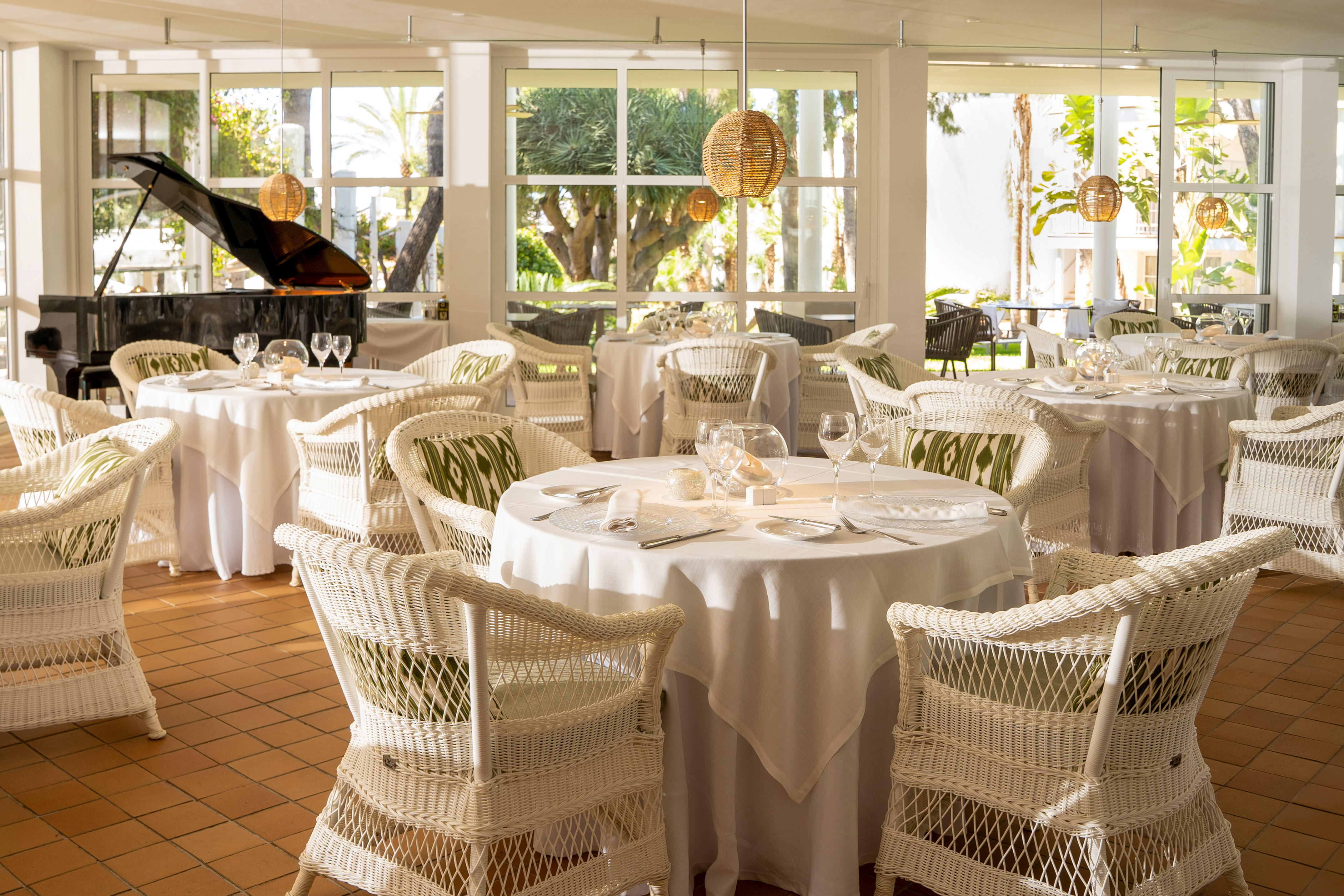 Bright restaurant with white wicker chairs, set tables, piano, and garden view.