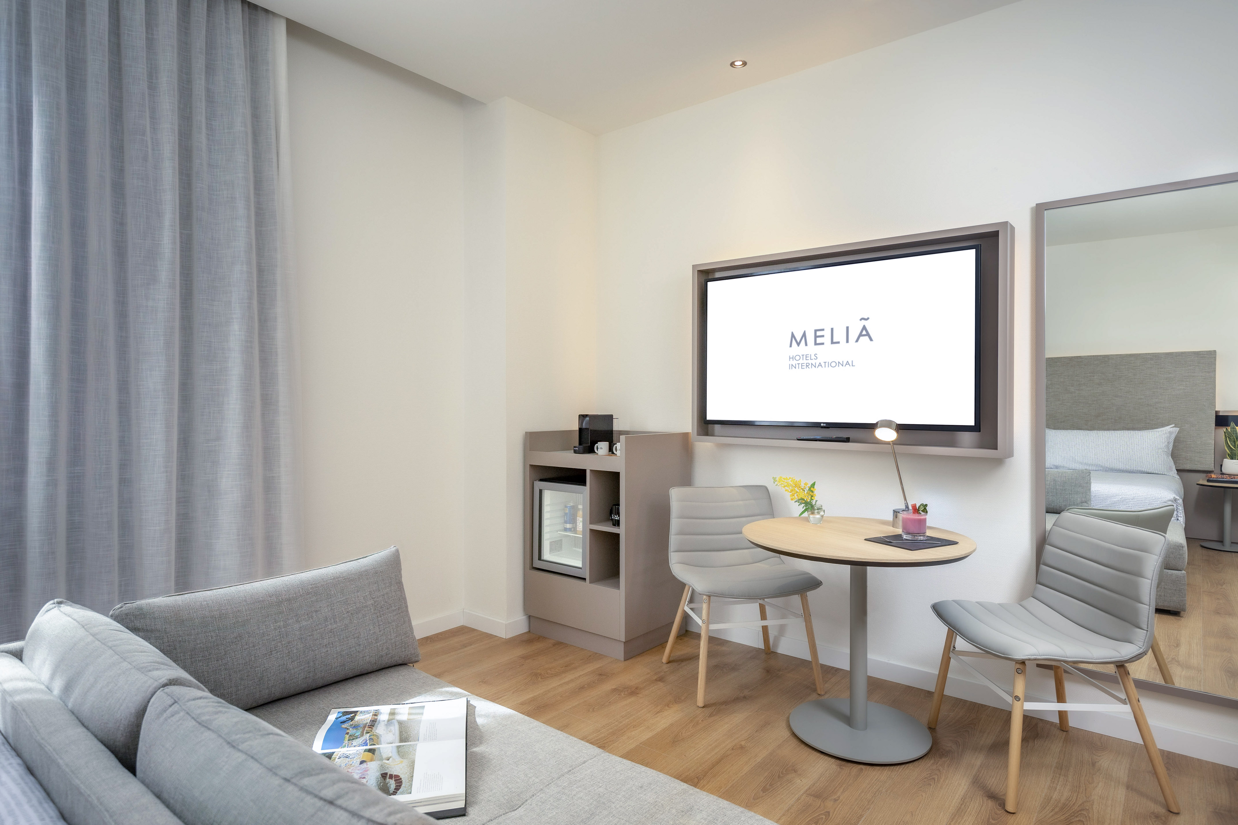 Modern Melia hotel suite: gray sofa, flat-screen TV, minibar, and dining area.