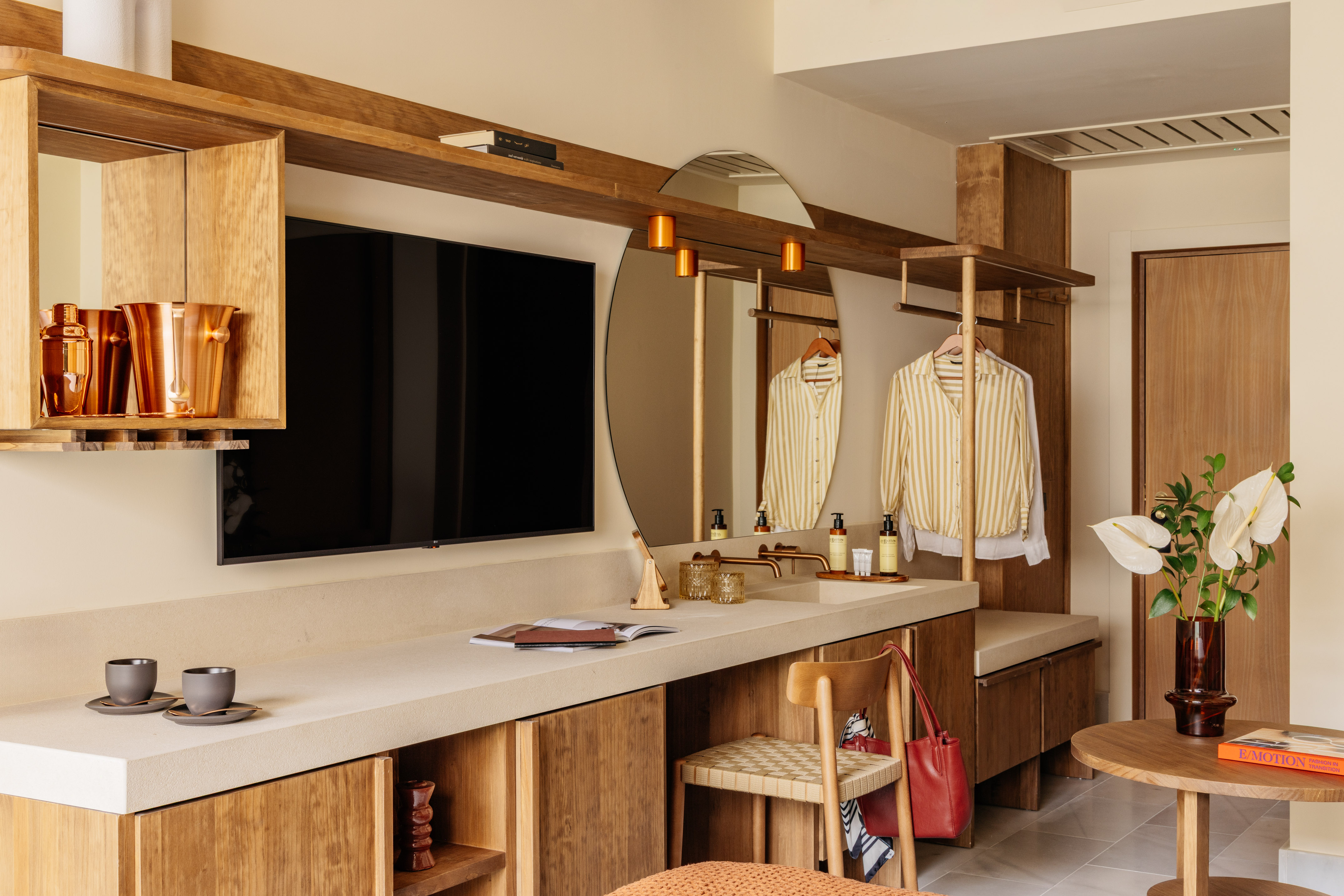 Modern hotel room with wooden vanity, integrated TV, round mirror, and open wardrobe.