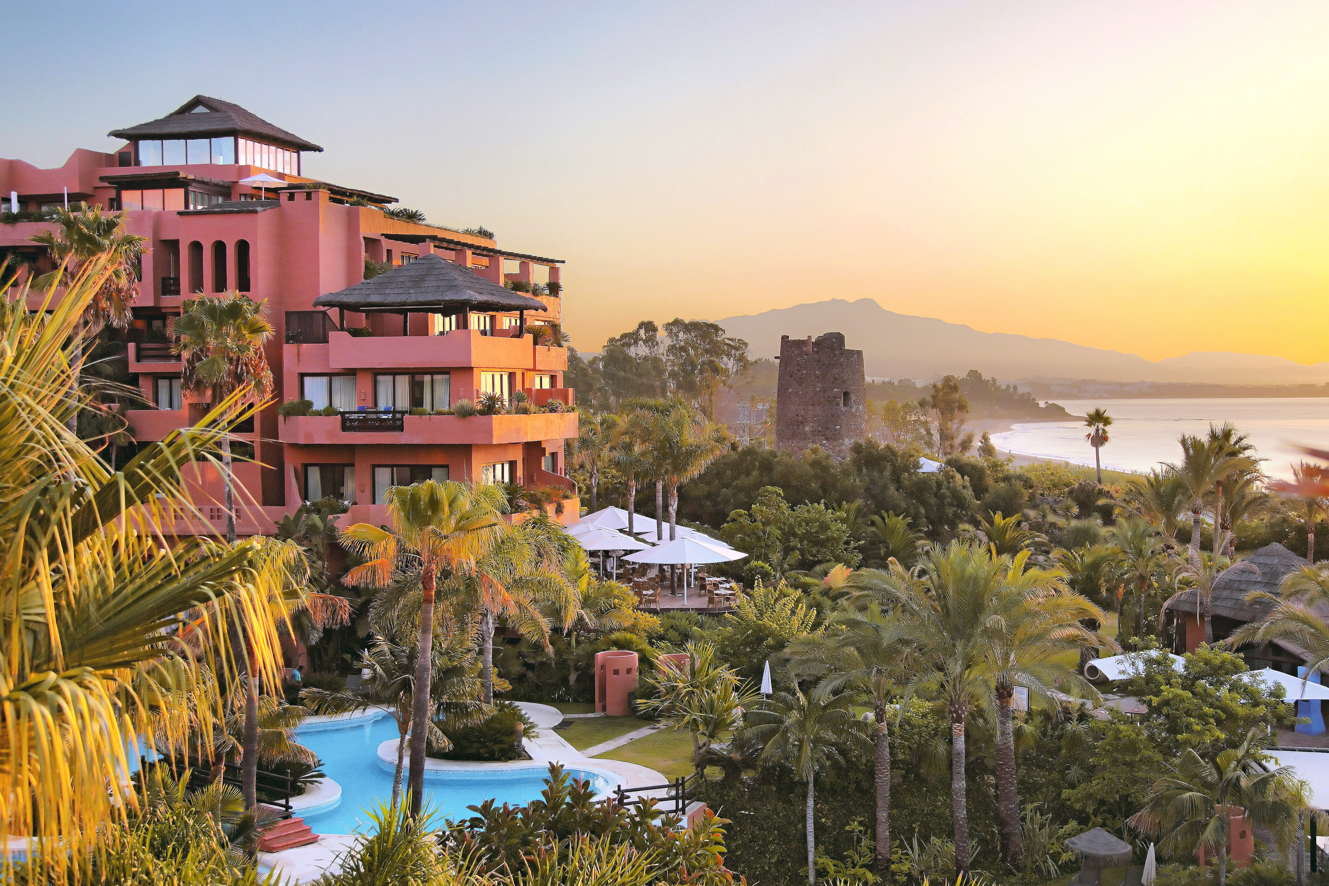 Coastal resort with pool, palm trees, historic tower, and sunset over the sea.