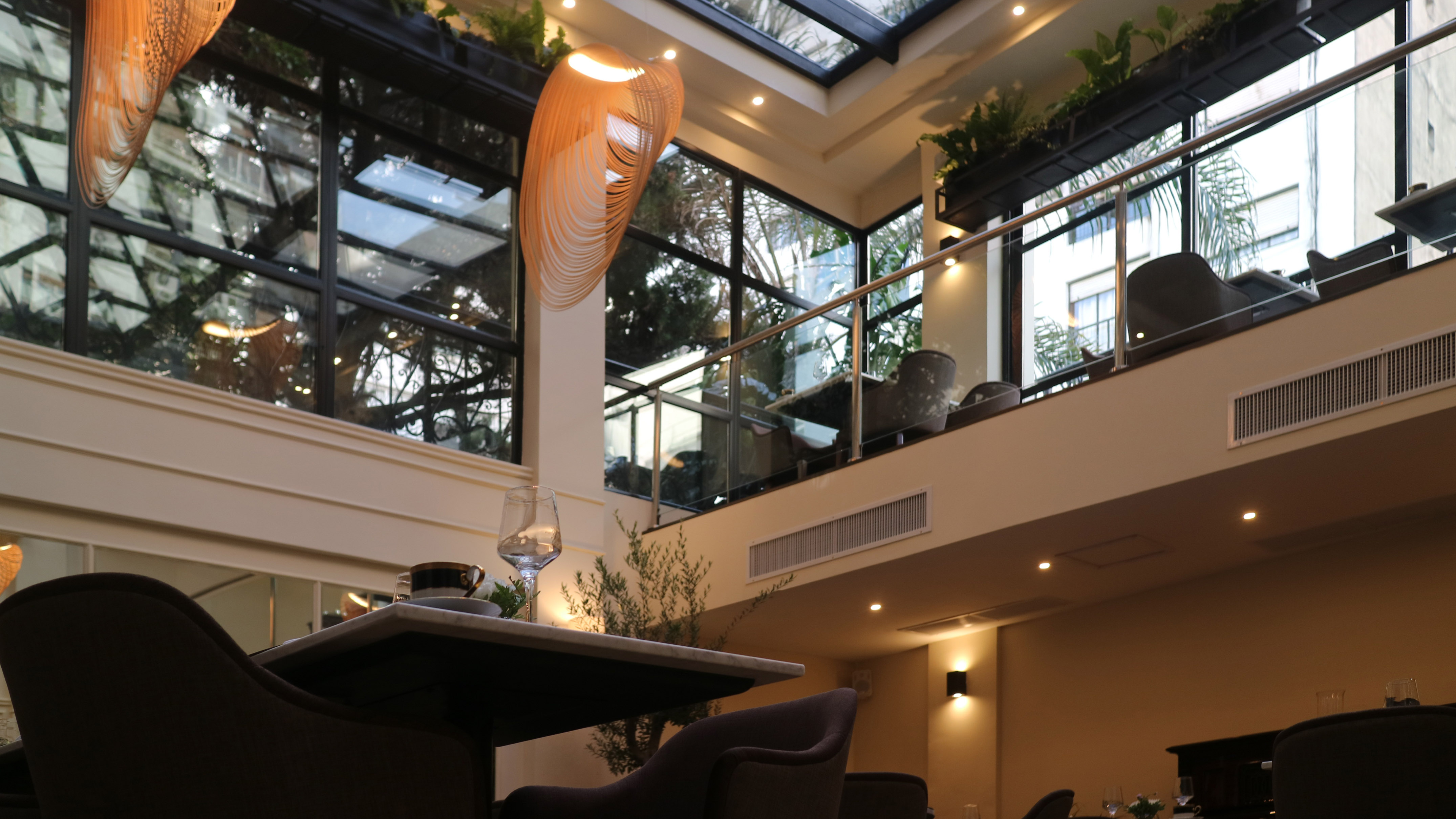 Modern two-story restaurant interior with glass roof, spiral chandelier, and plants.