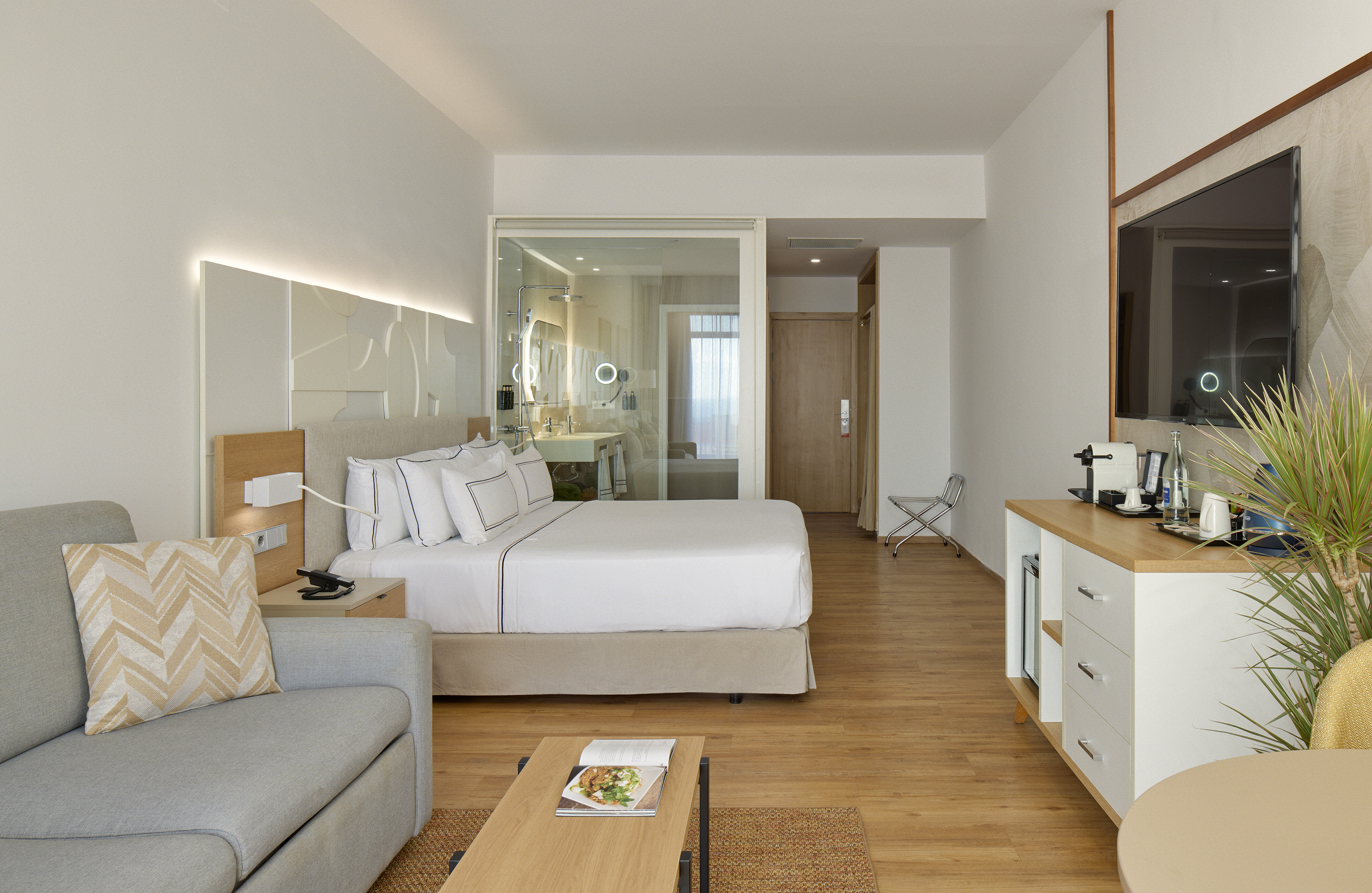 Modern hotel suite with bed, sofa, flat-screen TV, and glass bathroom.