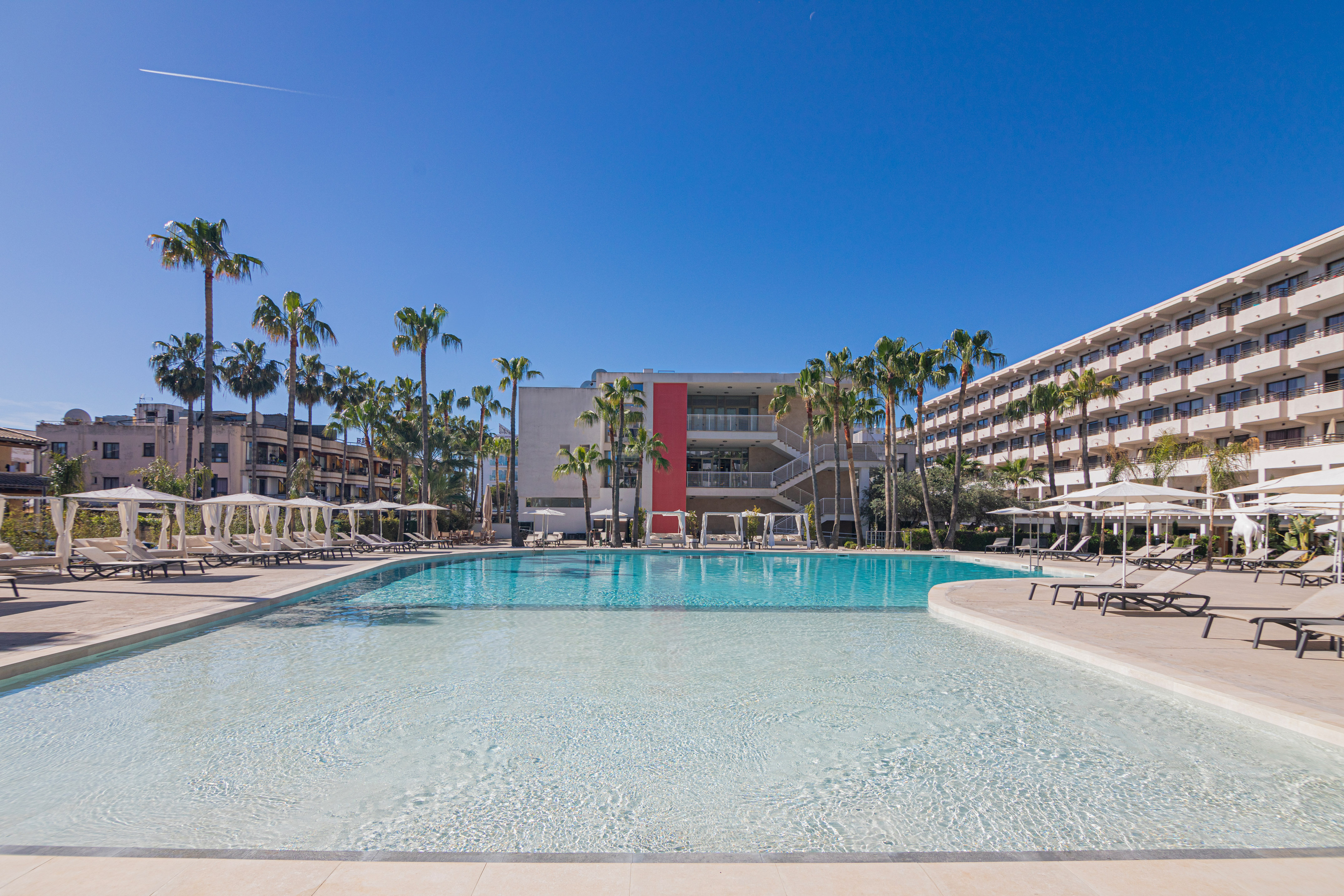 Sunny hotel resort with large outdoor pool, palm trees, and lounge chairs.