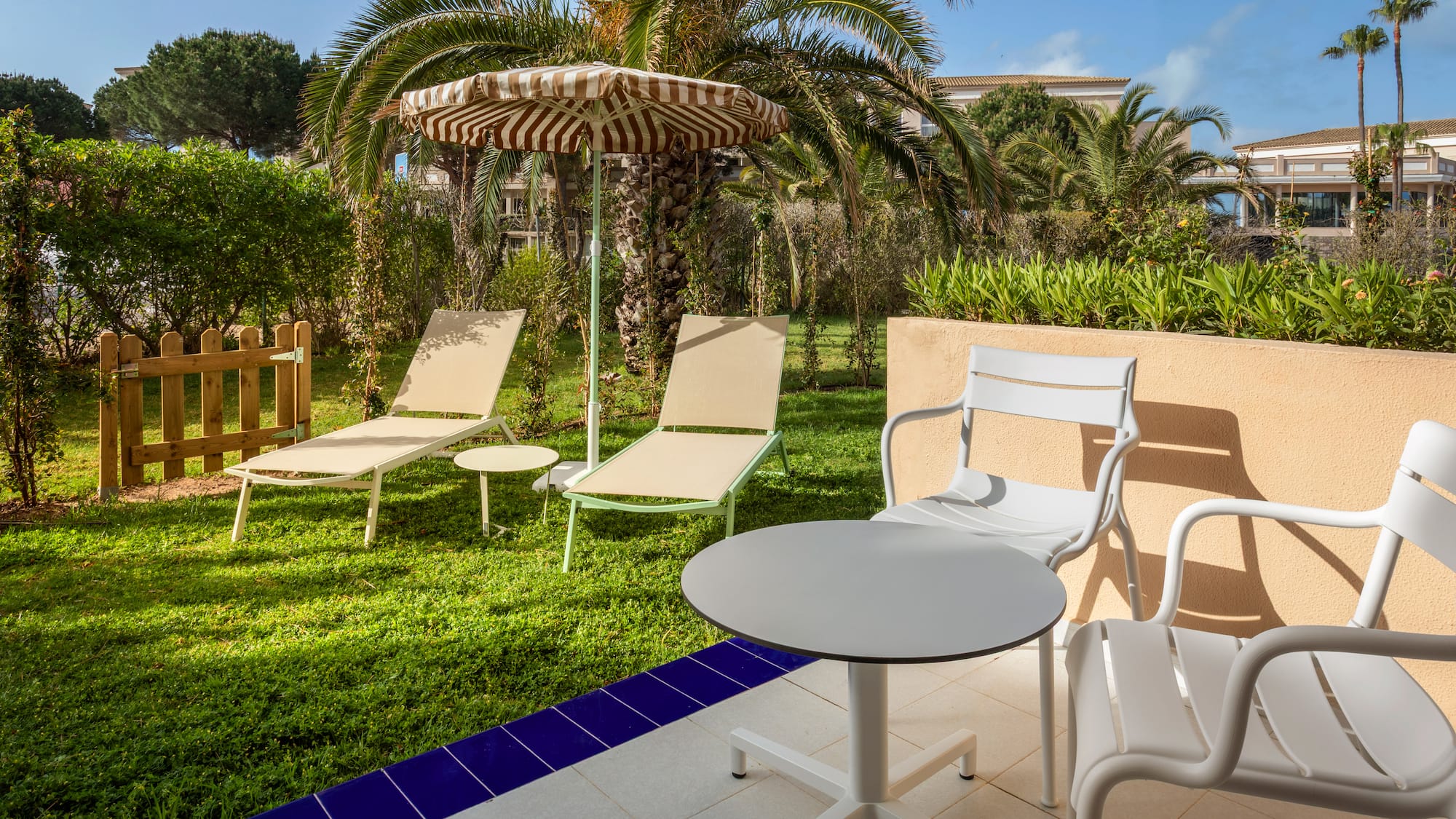 Sunny hotel patio with garden, lounge chairs, striped umbrella, and palm trees.