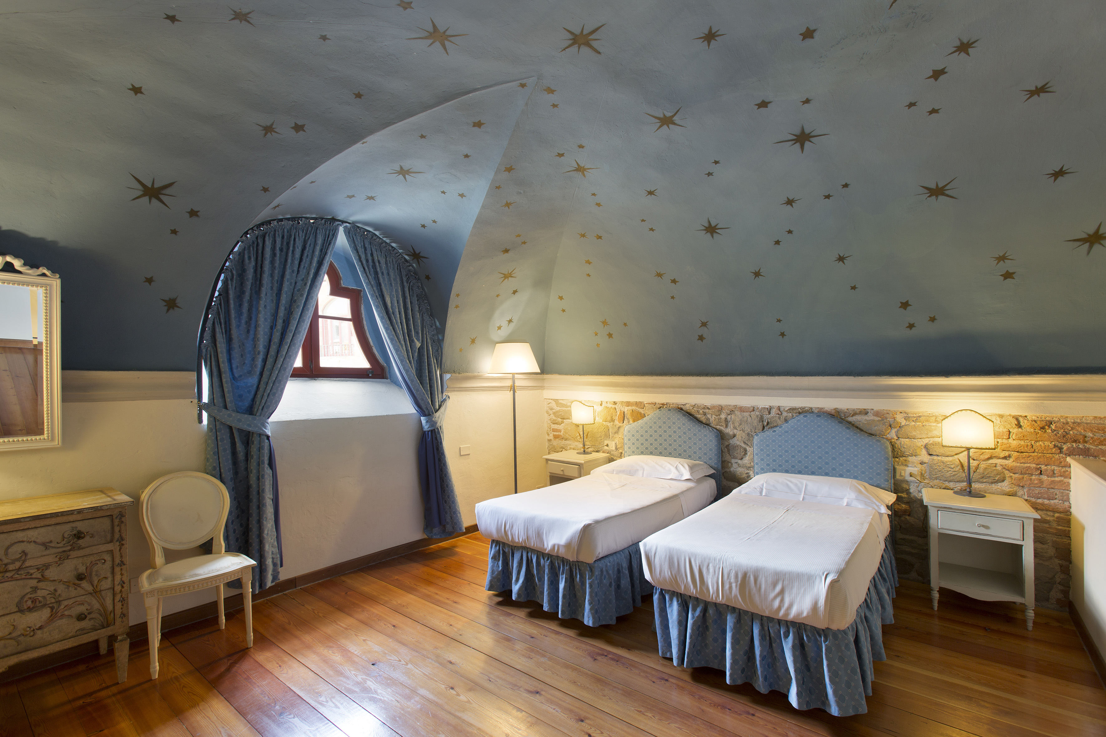 Hotel bedroom with twin beds, star-painted arched ceiling, and wooden floor.