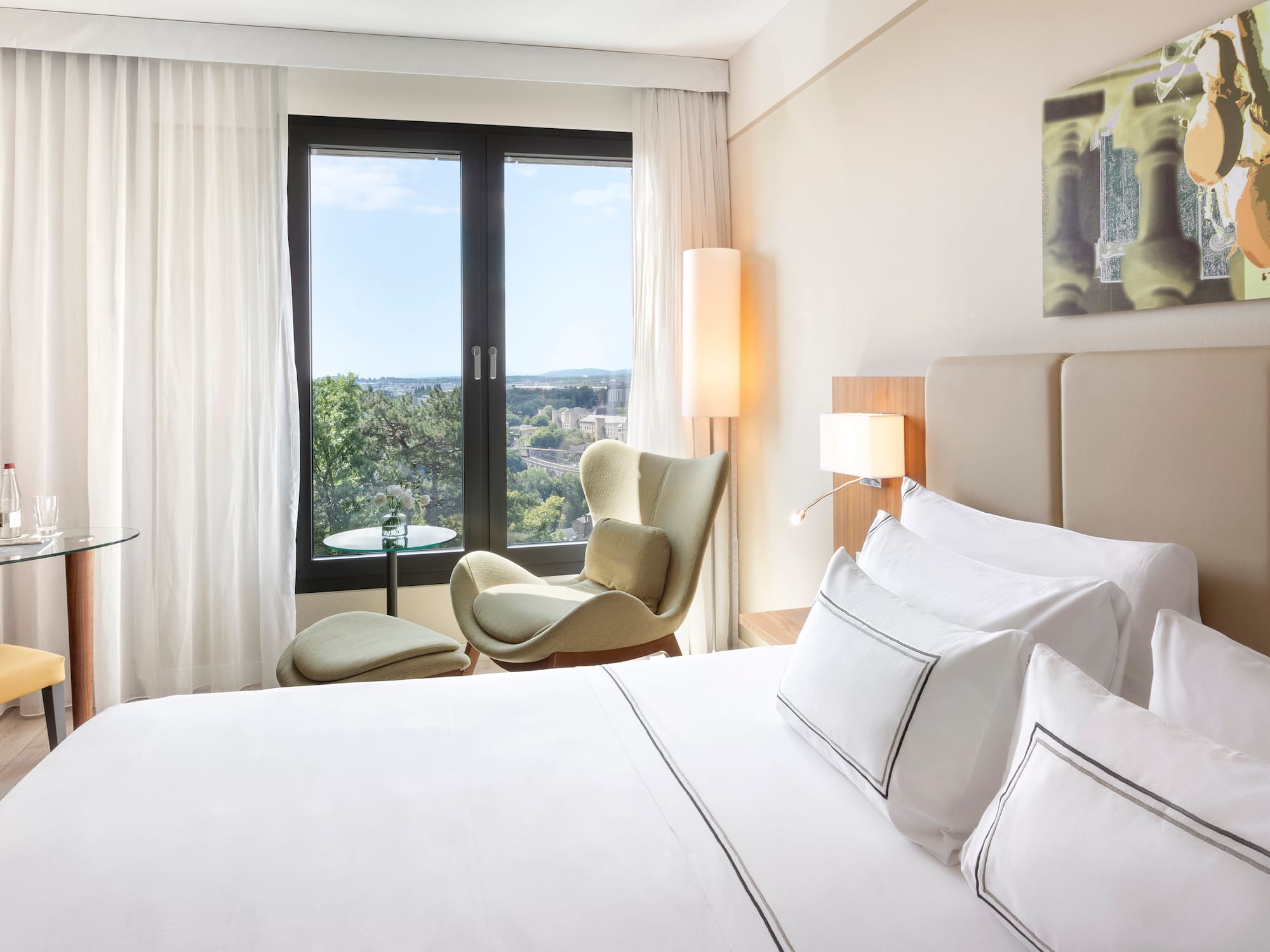 Bright hotel room with city view, white bed, green armchair, and modern decor.