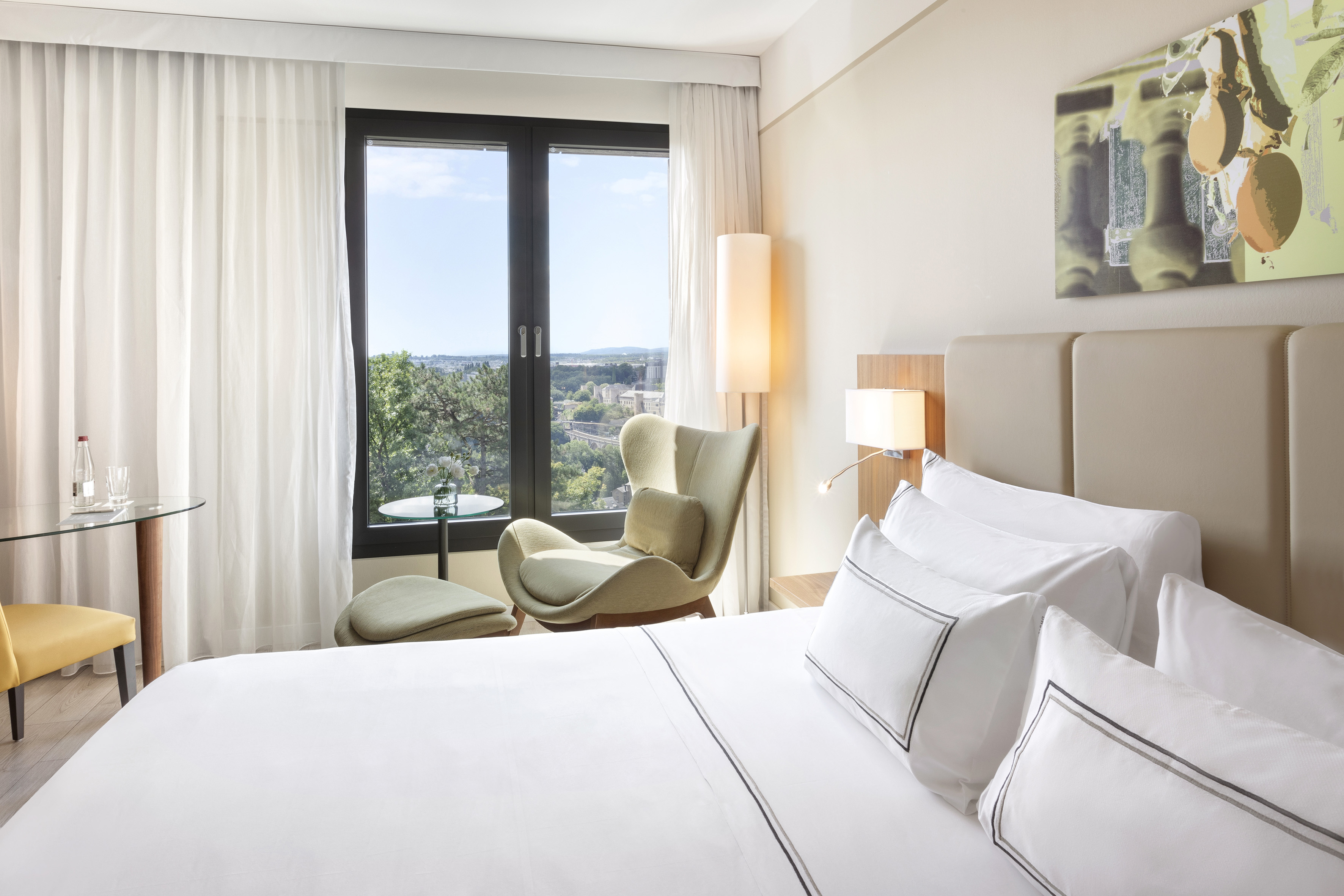Bright hotel room with city view, white bed, green armchair, and modern decor.