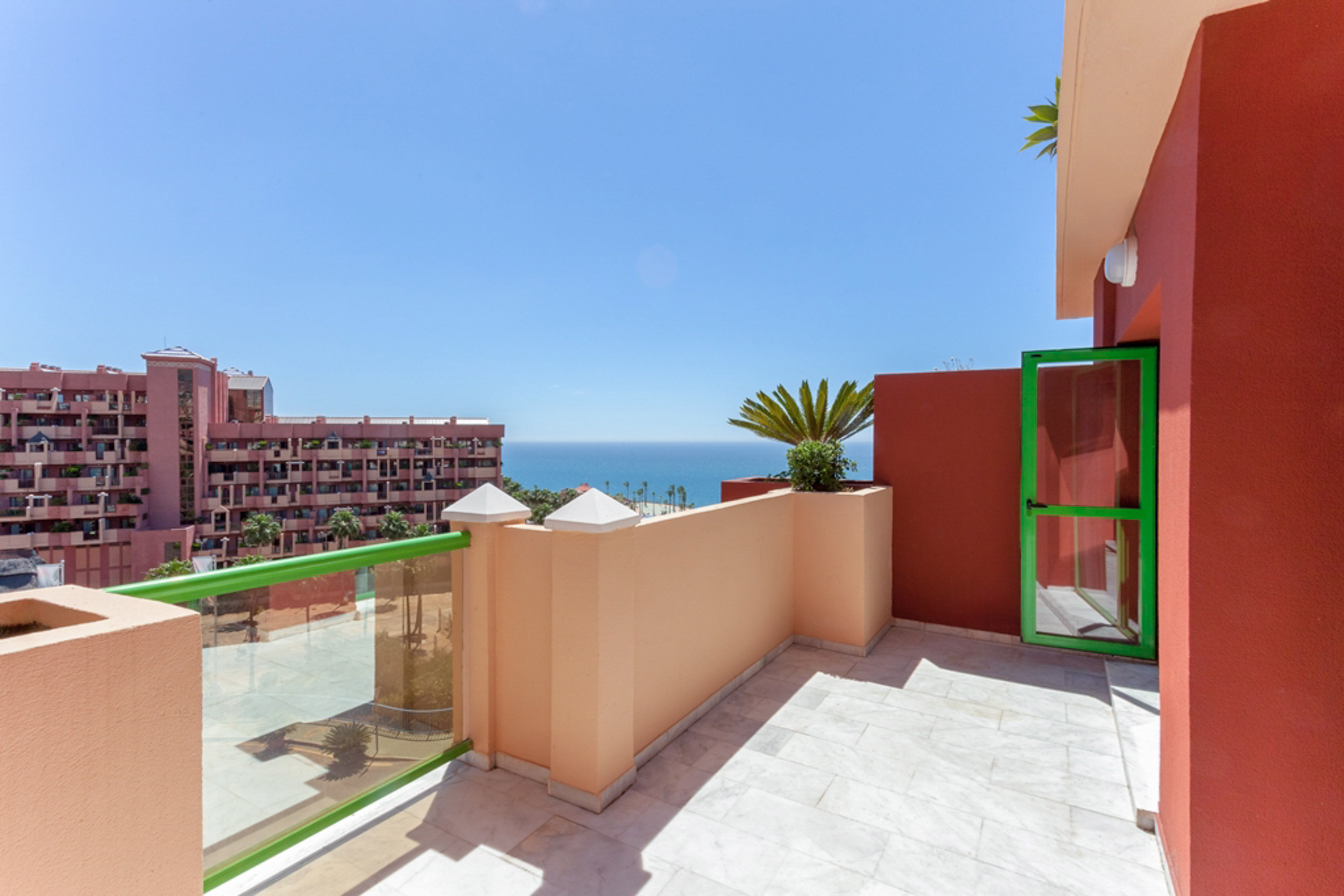 Sunny balcony with ocean view, green railing, marble floor, and terracotta building.