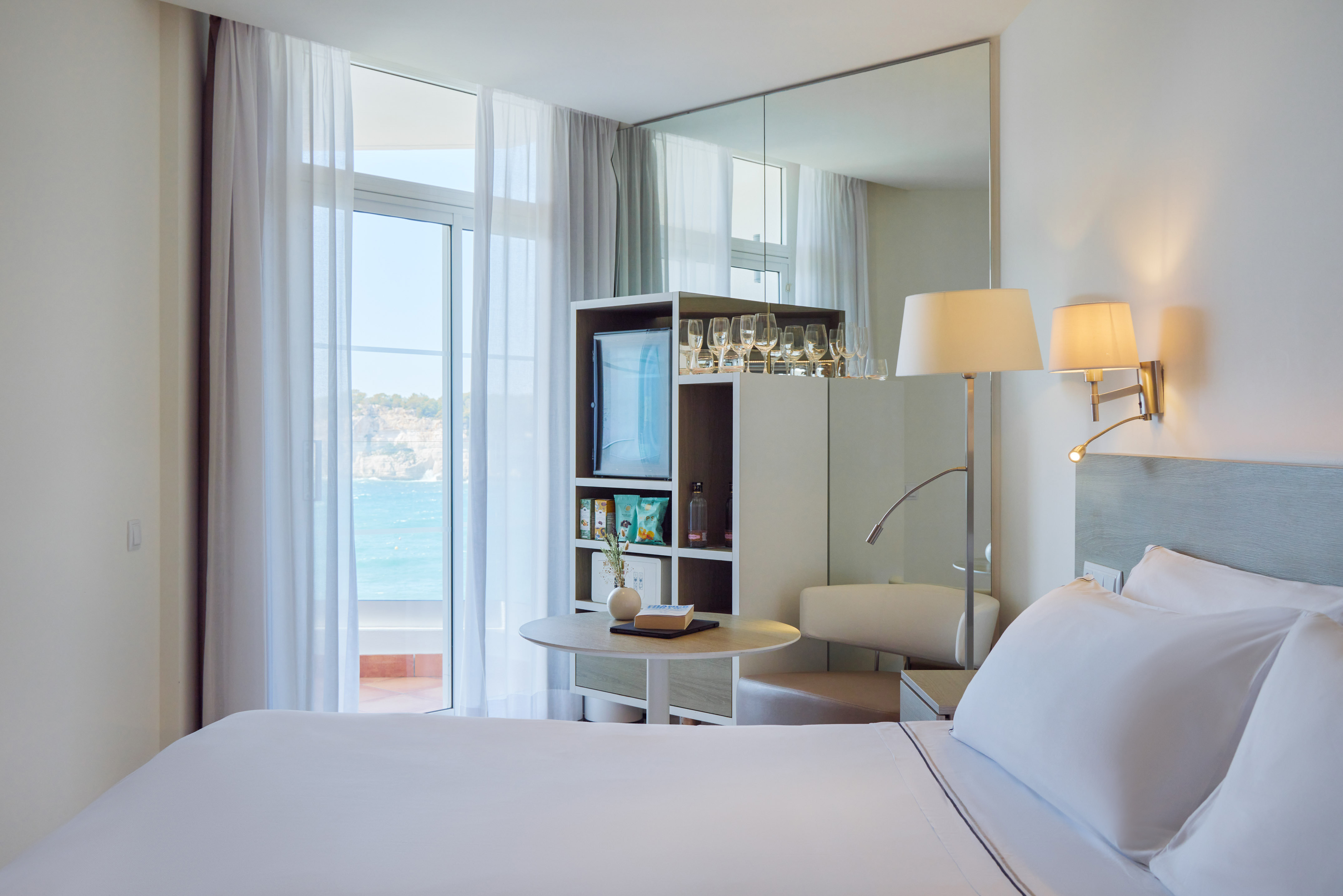 Modern hotel room with ocean view balcony, white bed, and contemporary minibar.