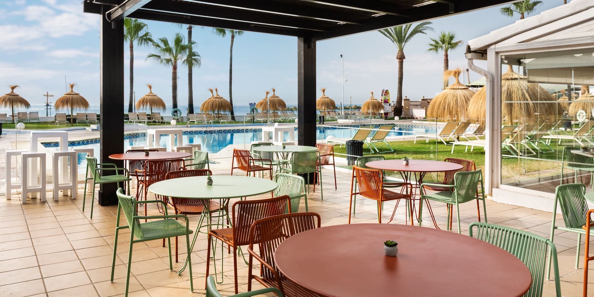 Terraza de hotel con mesas, sillas, piscina, palmeras y playa.