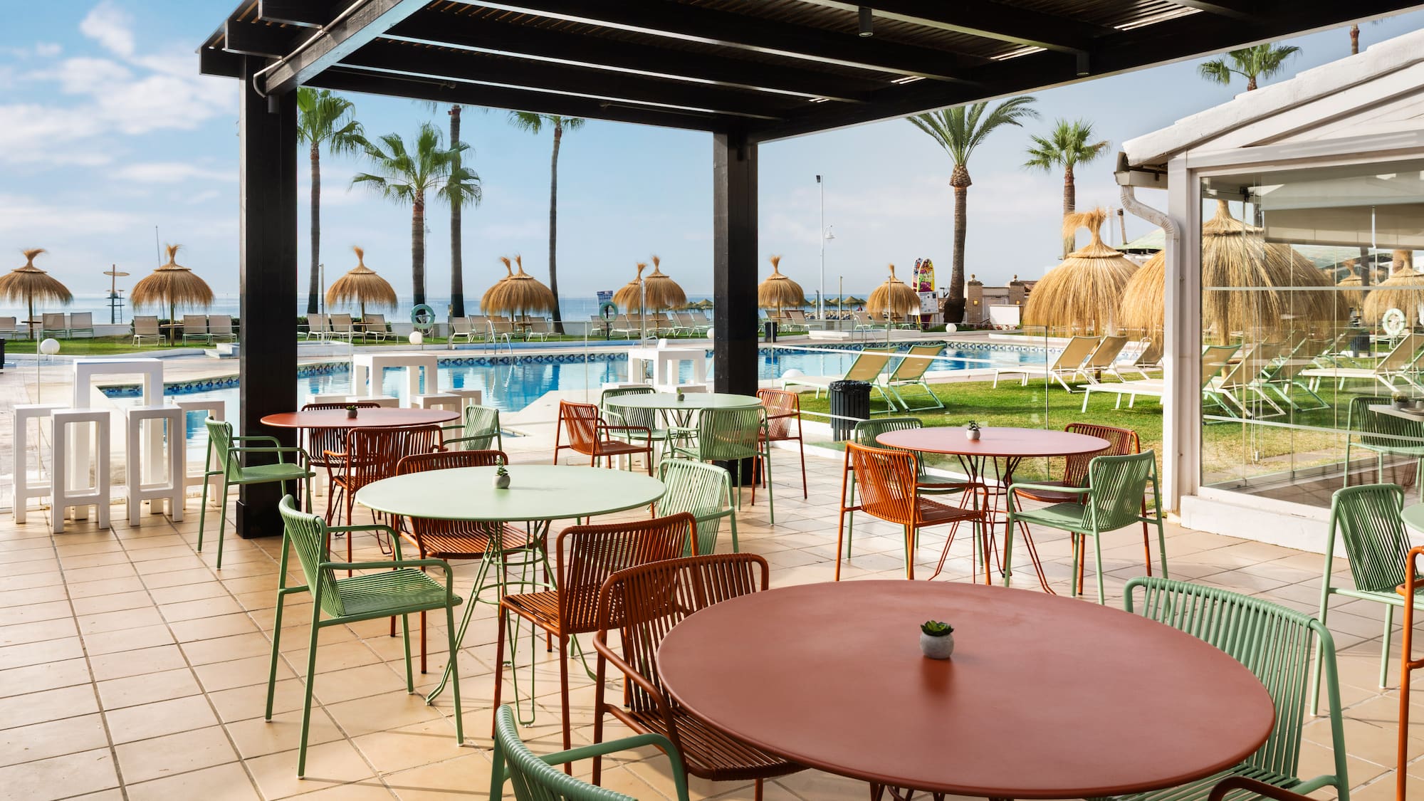 Terraza de hotel con mesas, sillas, piscina, palmeras y playa.