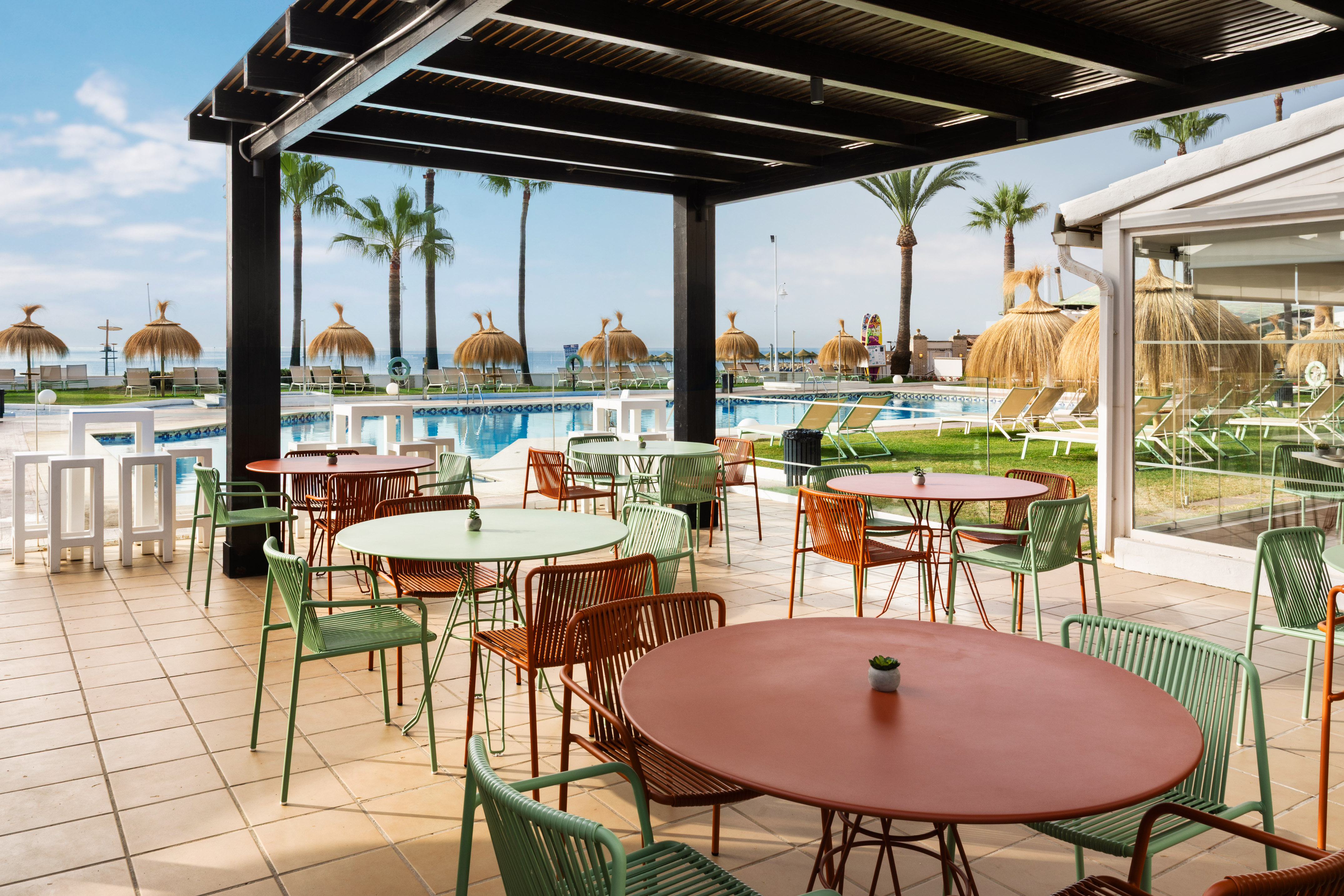 Terraza de hotel con mesas, sillas, piscina, palmeras y playa.