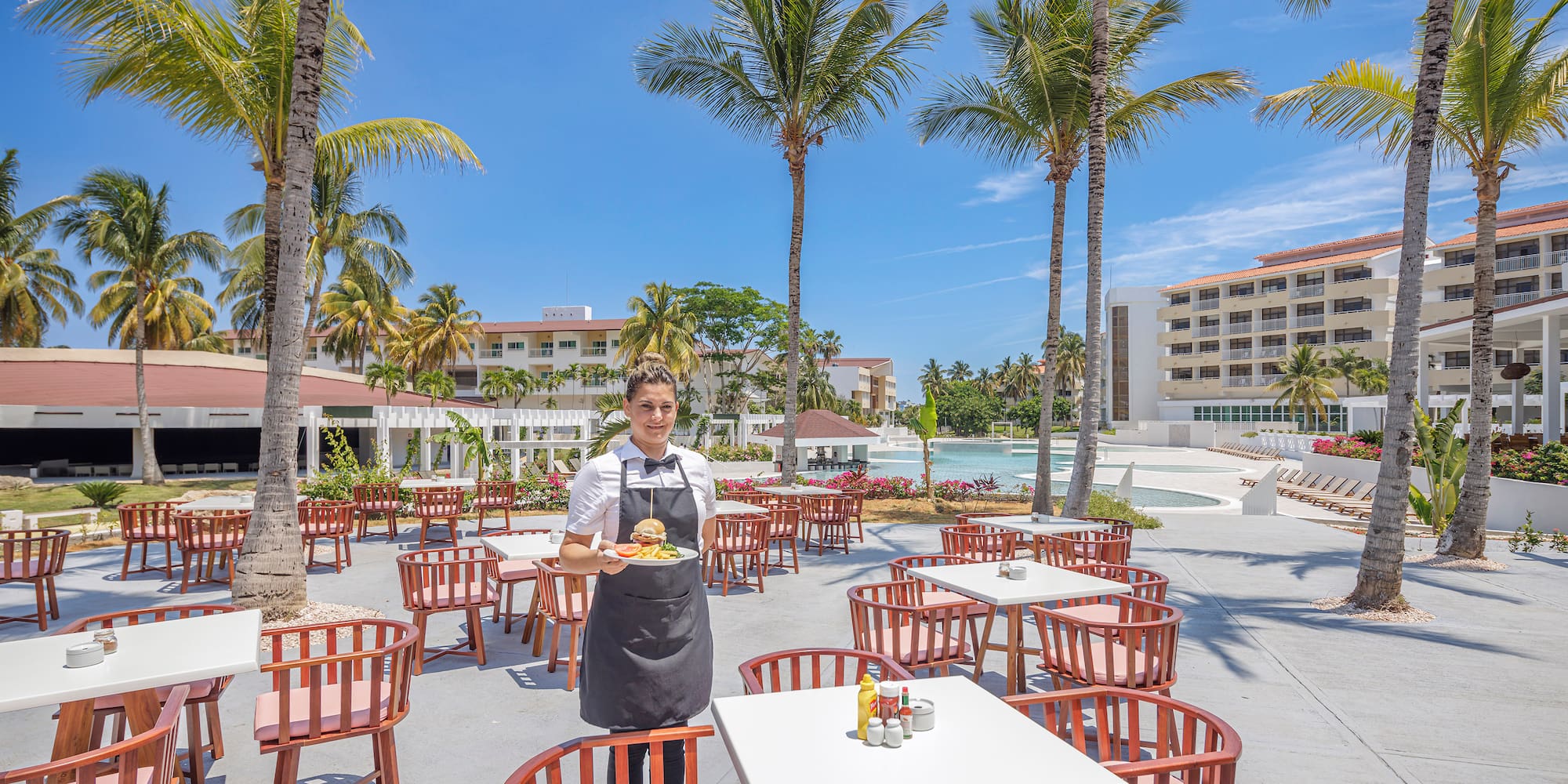 a person in a black apron standing in a courtyard with tables and chairs