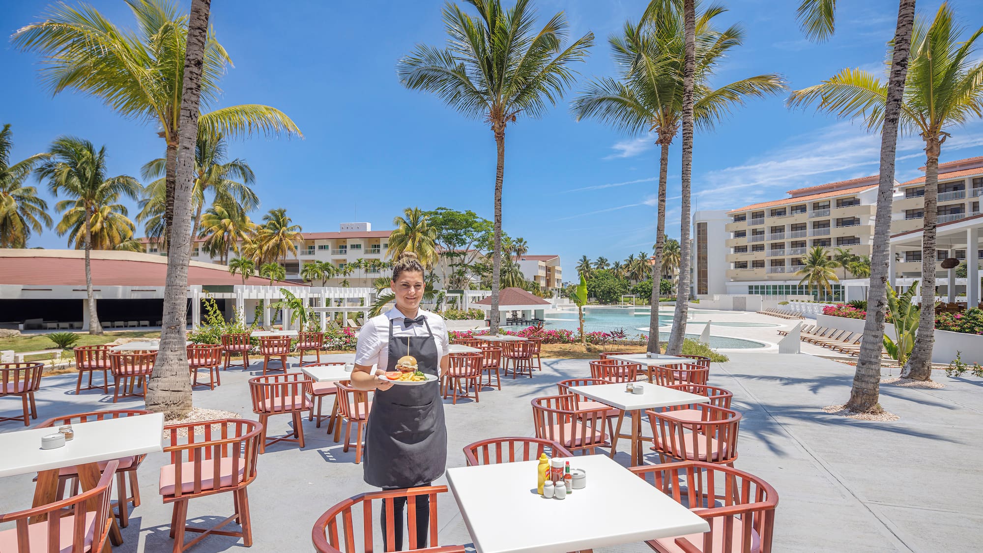 a person in a black apron standing in a courtyard with tables and chairs