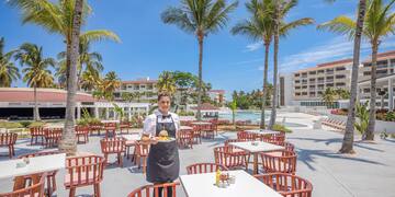 a person in a black apron standing in a courtyard with tables and chairs