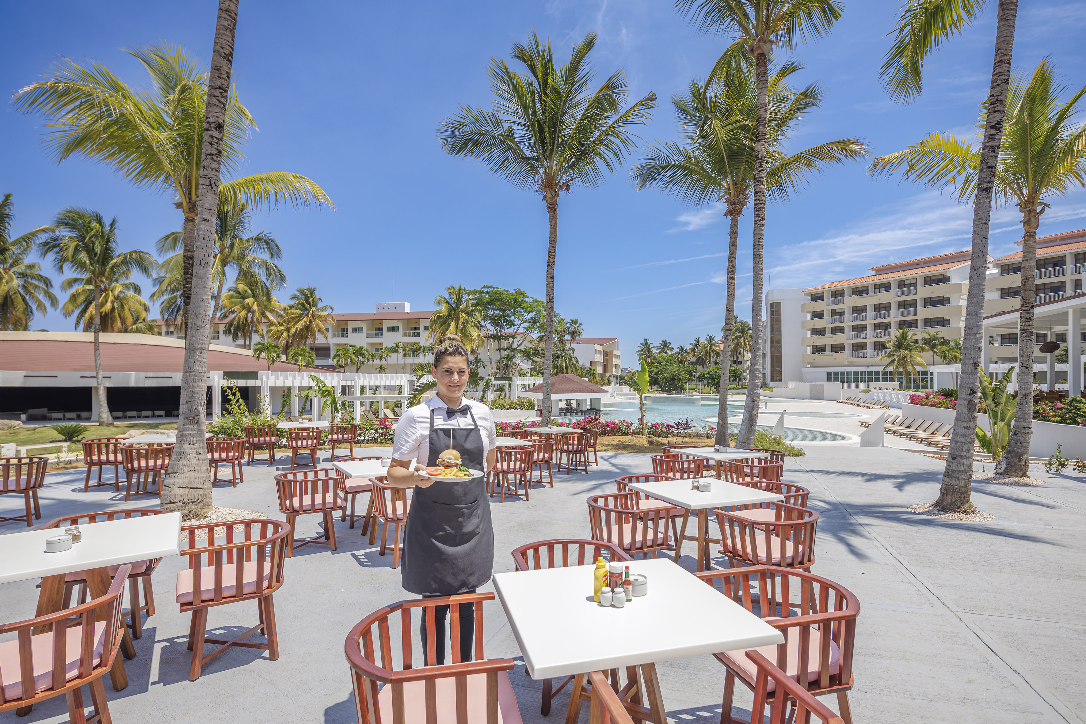 a person in a black apron standing in a courtyard with tables and chairs