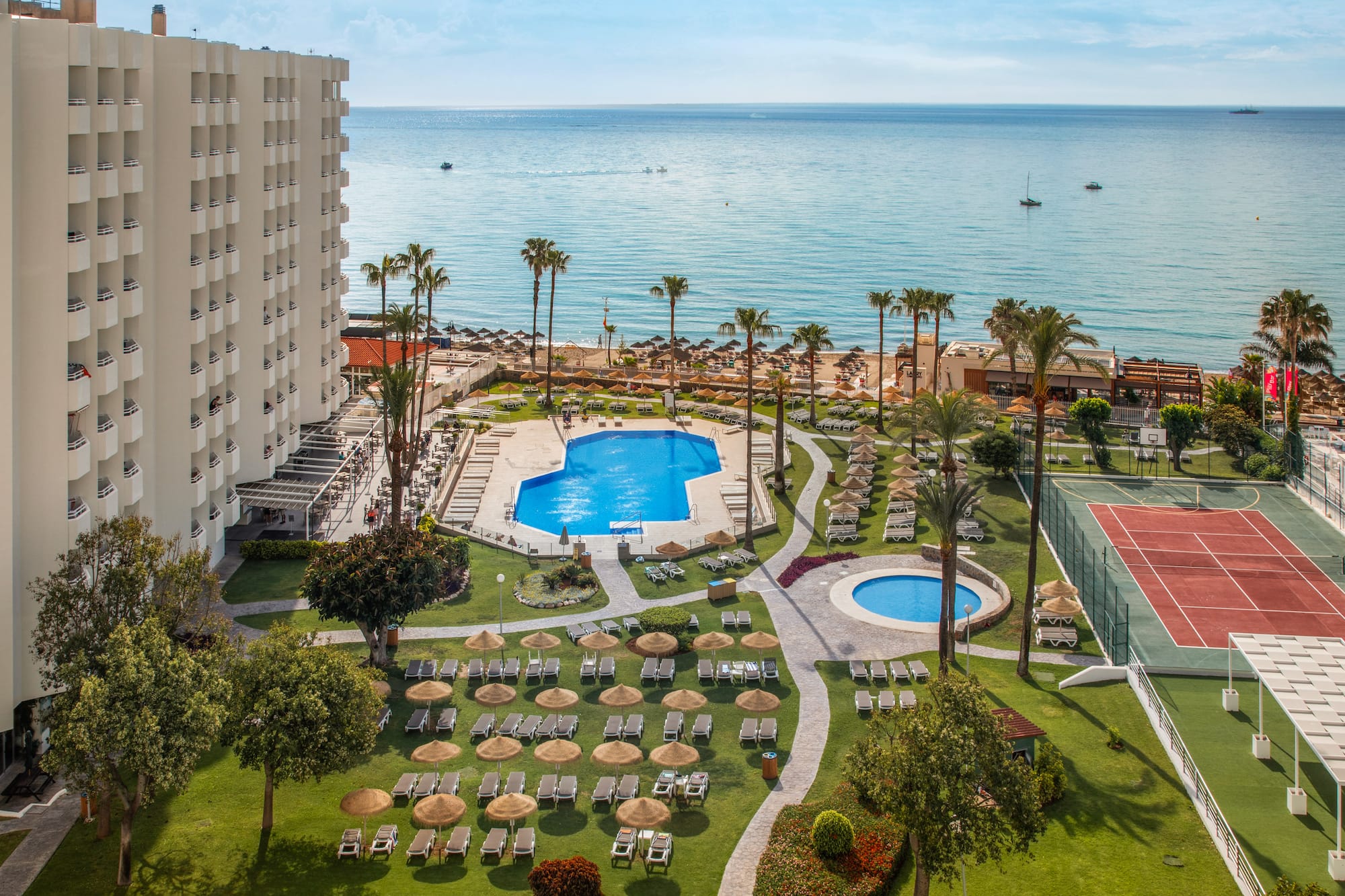Oceanfront resort with hotel, pools, sunny beach, palm trees, and tennis court.