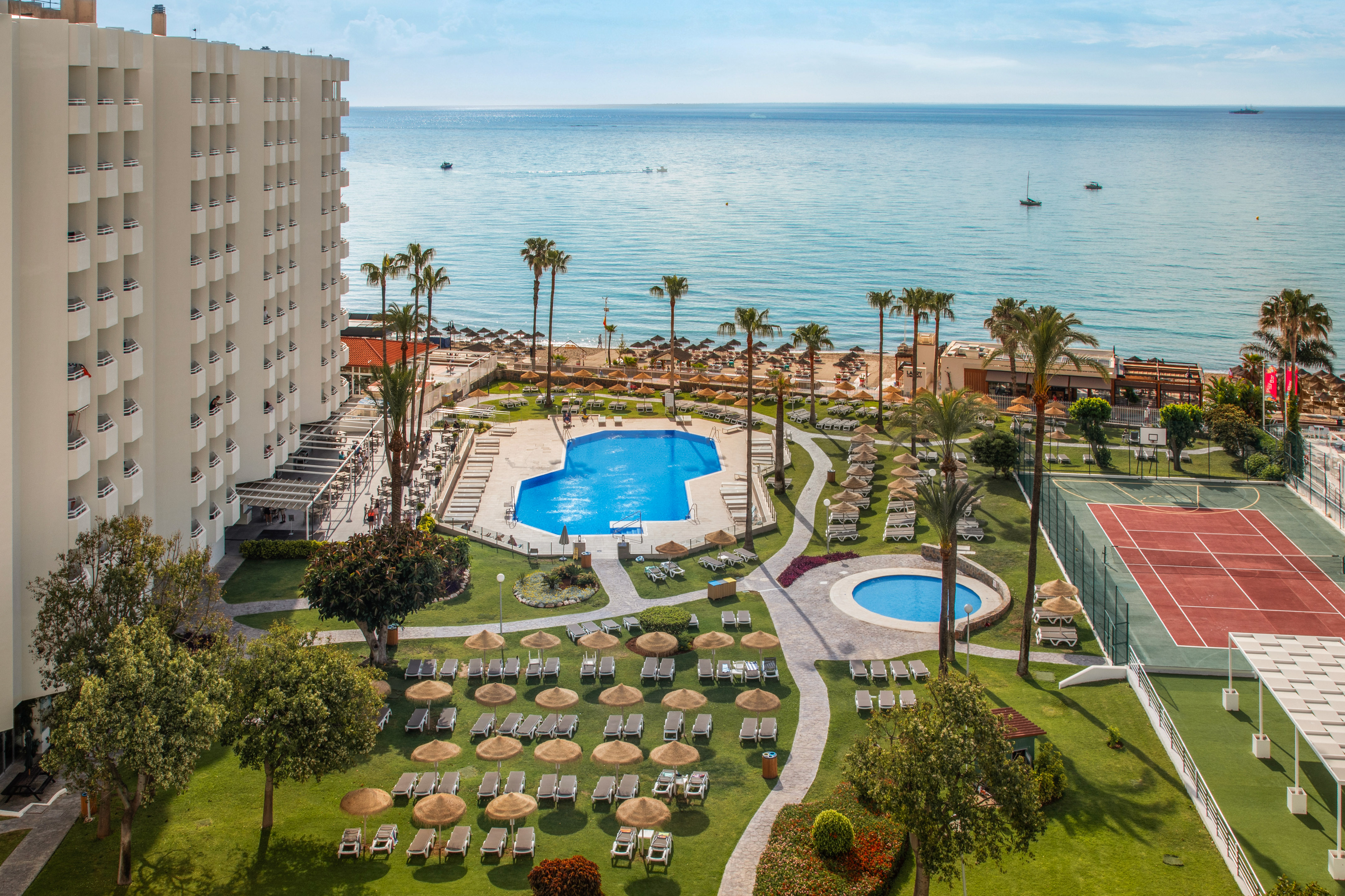 Oceanfront resort with hotel, pools, sunny beach, palm trees, and tennis court.