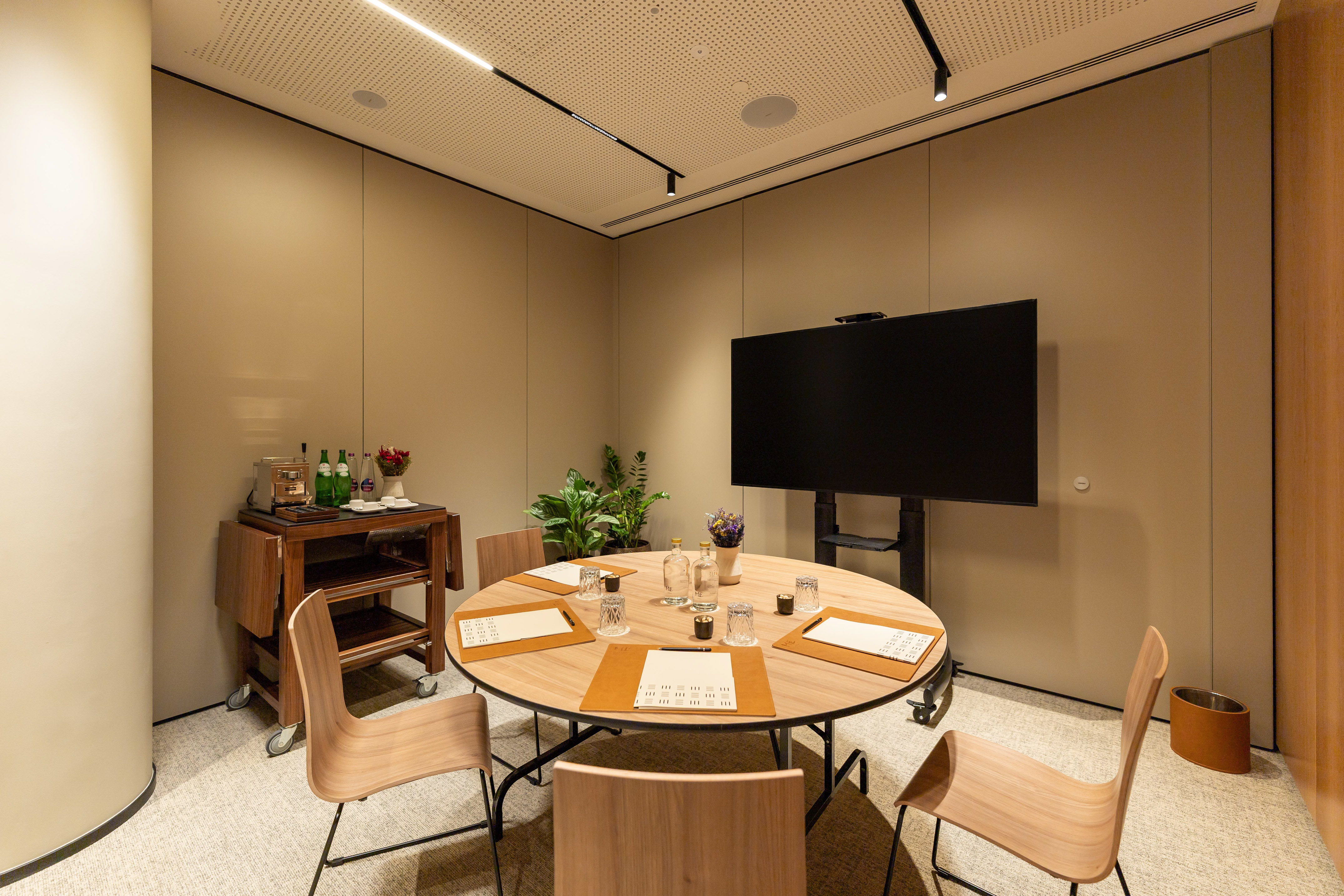 Modern meeting room with round table, chairs, large screen, and service cart.