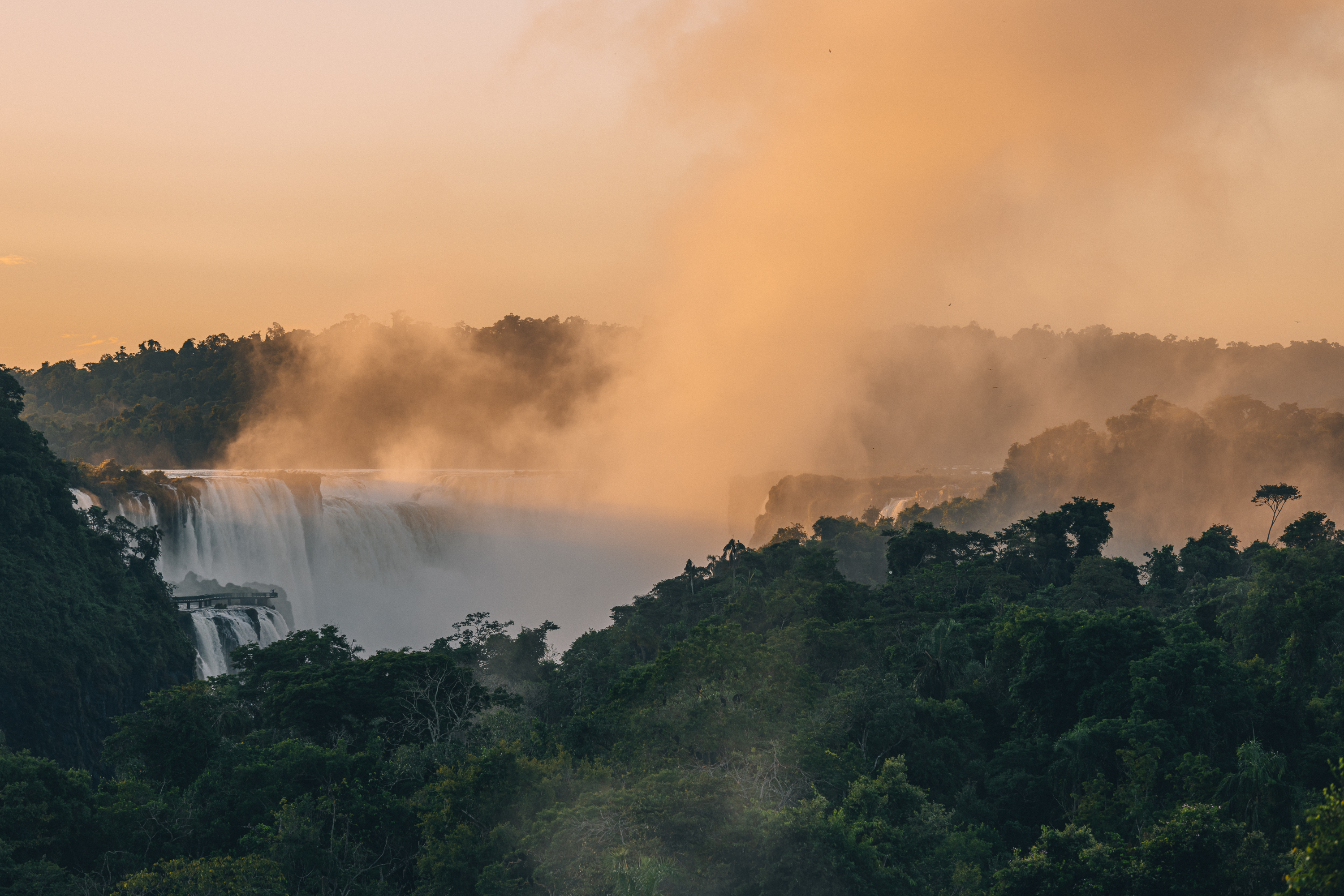Misty cascades of Iguazu Falls at sunset, enveloped by dense jungle.