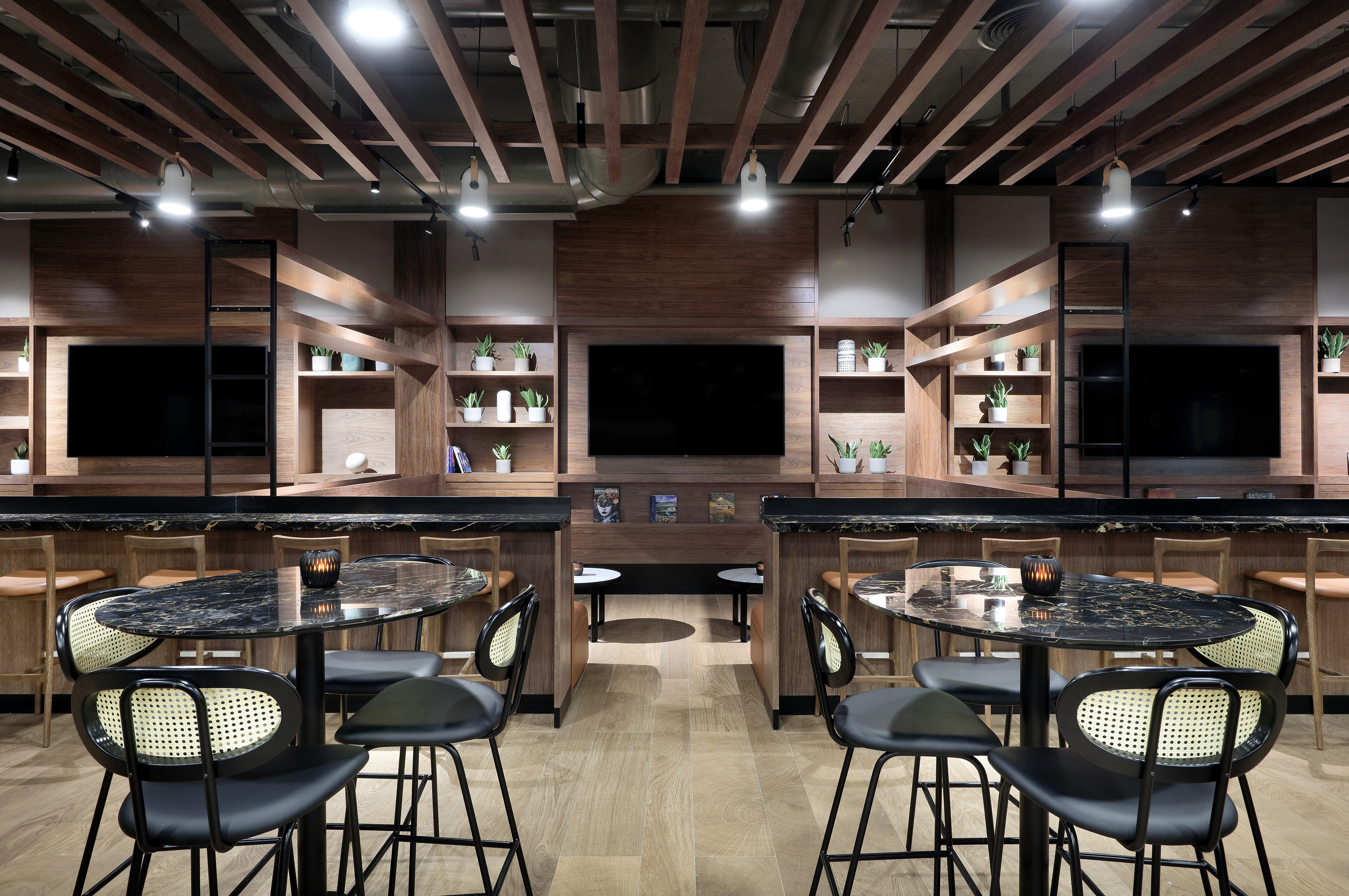 Modern lounge interior, wood panels, TVs, plants, black marble tables, bar seating.