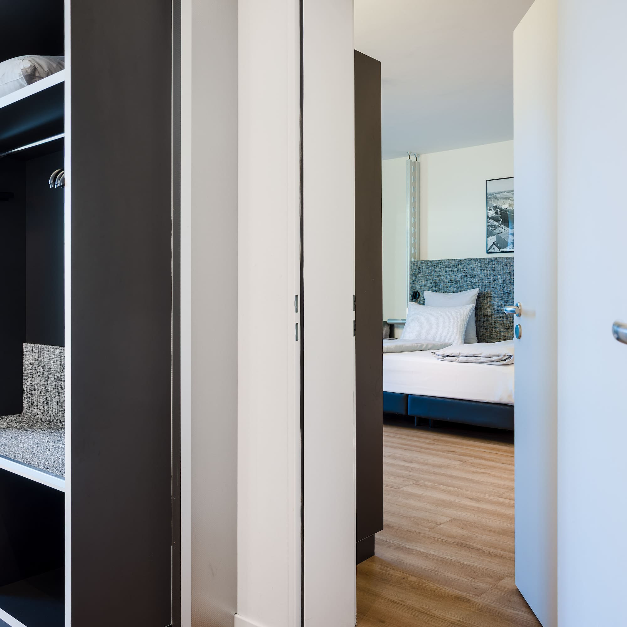 Modern hotel room entrance, open wardrobe with storage, glimpse of bedroom.