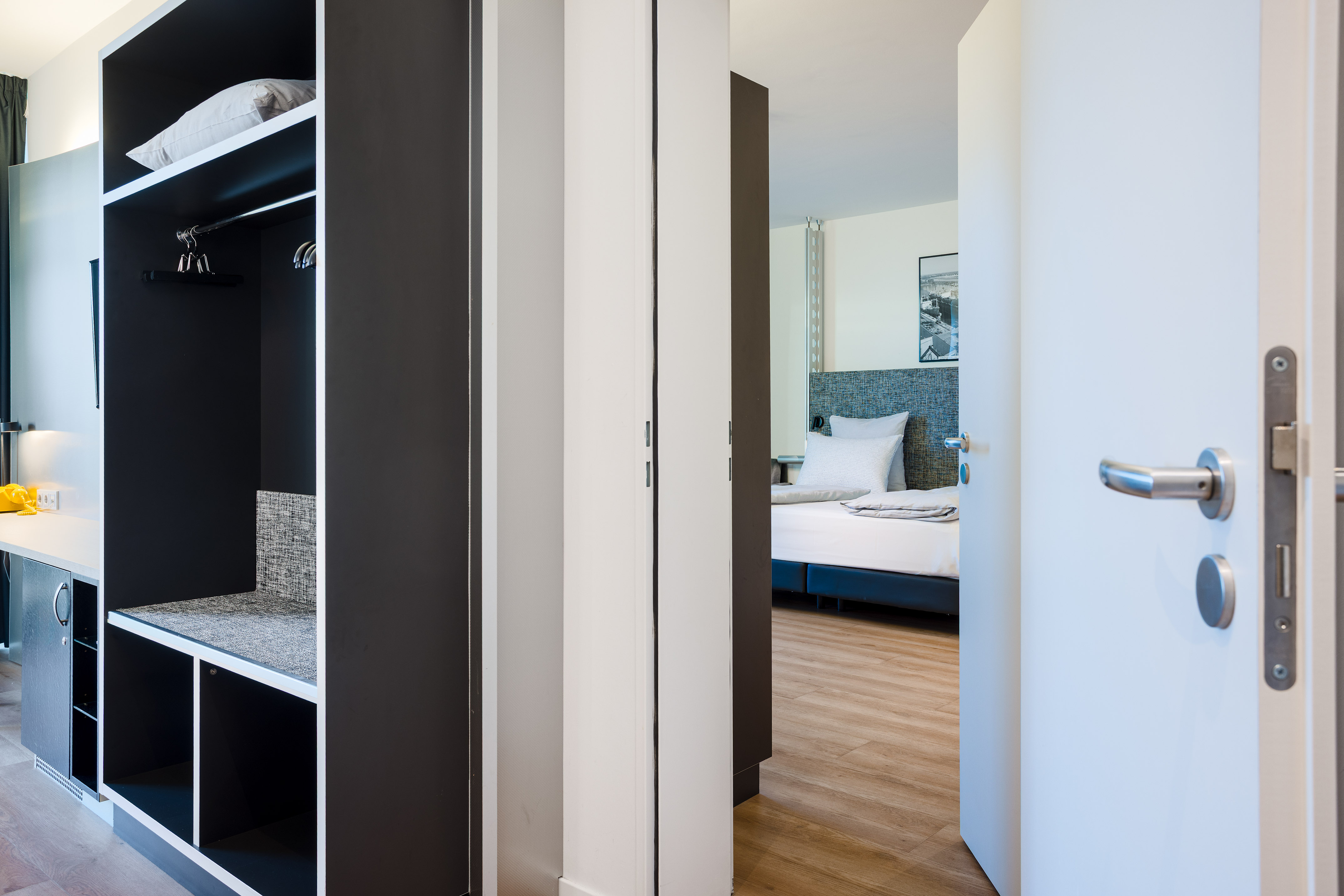 Modern hotel room entrance, open wardrobe with storage, glimpse of bedroom.