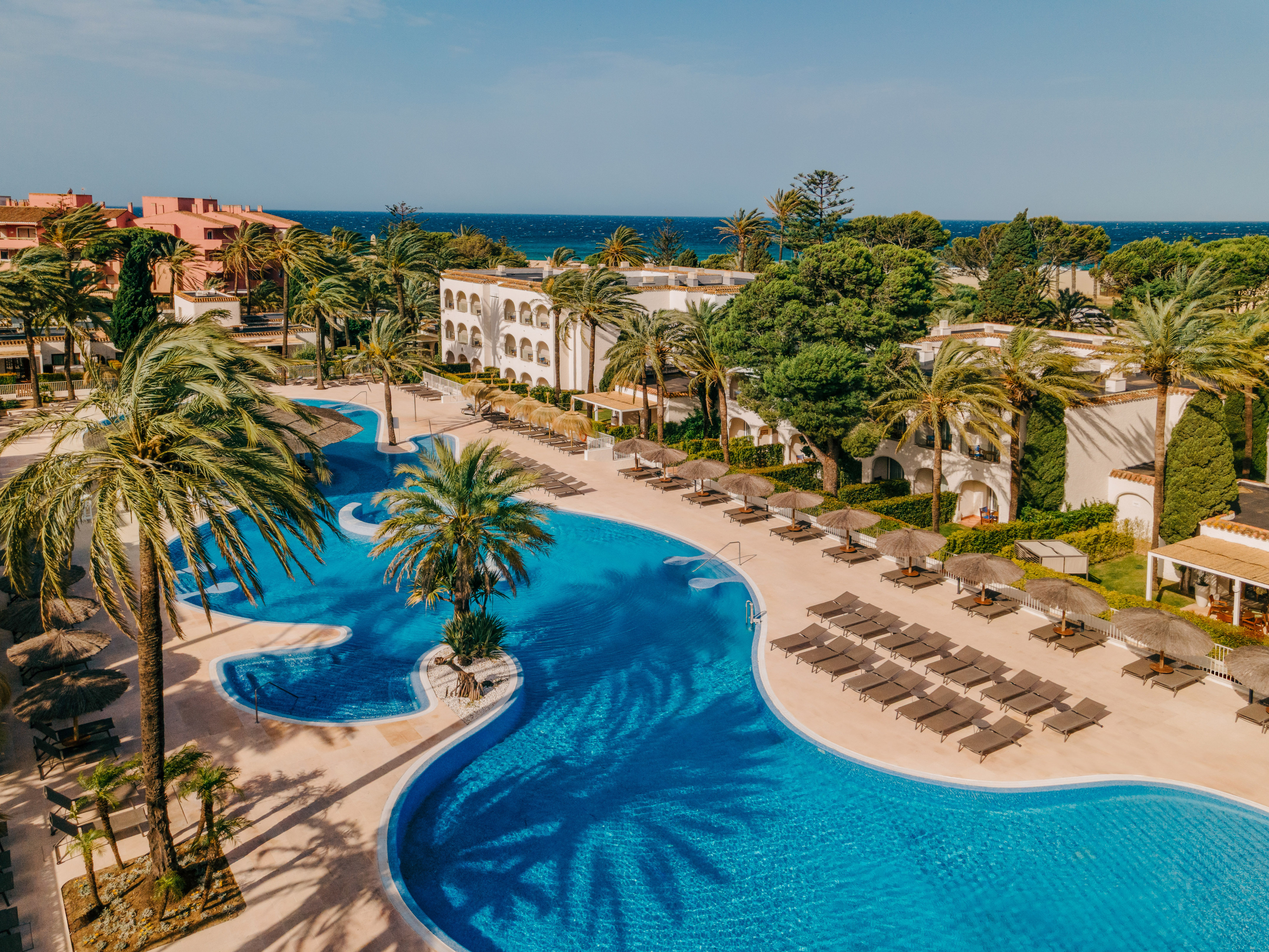 Tropical resort aerial: large pool, palm trees, hotel, ocean view.