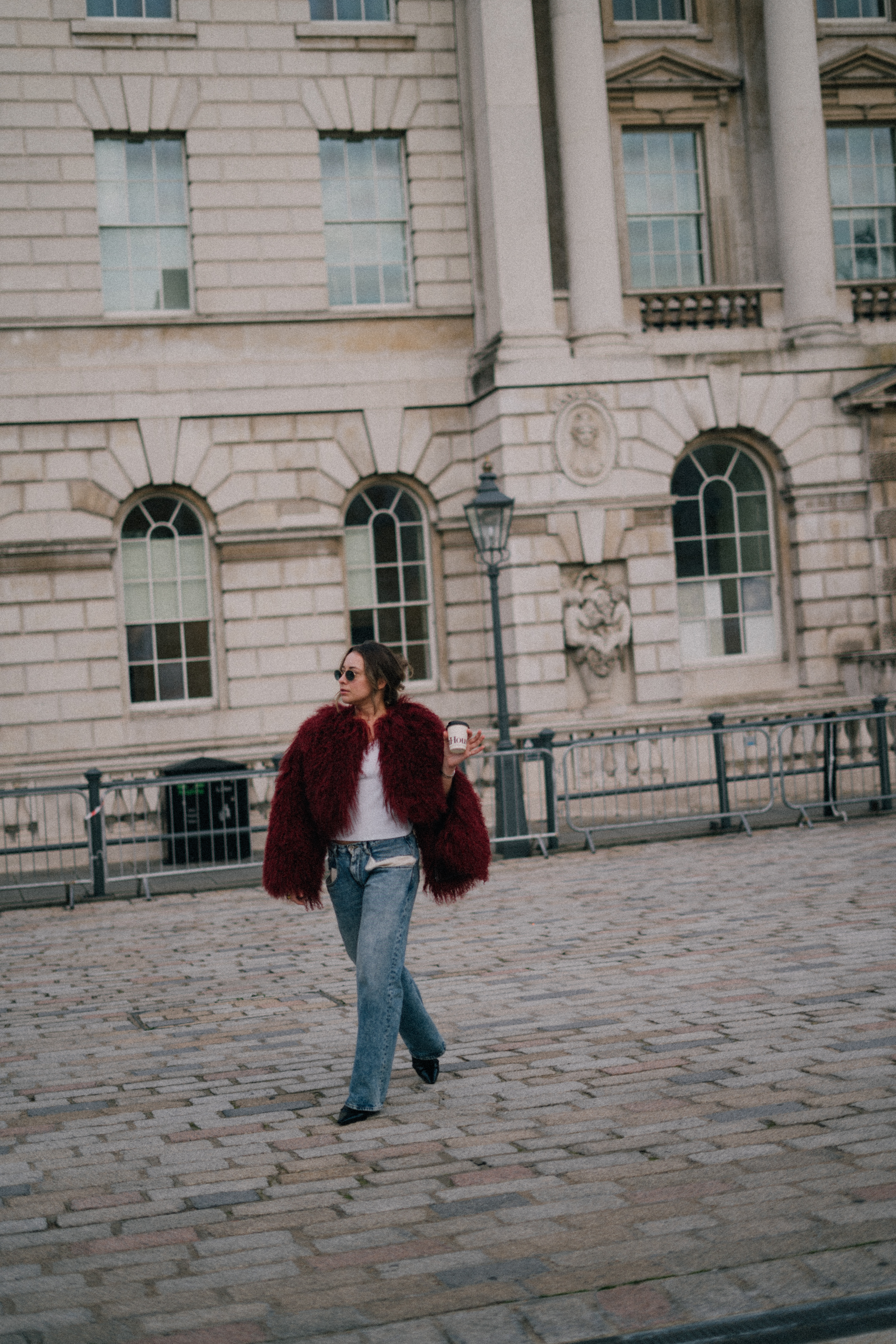 Woman in maroon faux fur jacket, jeans, walking on cobblestone by building.