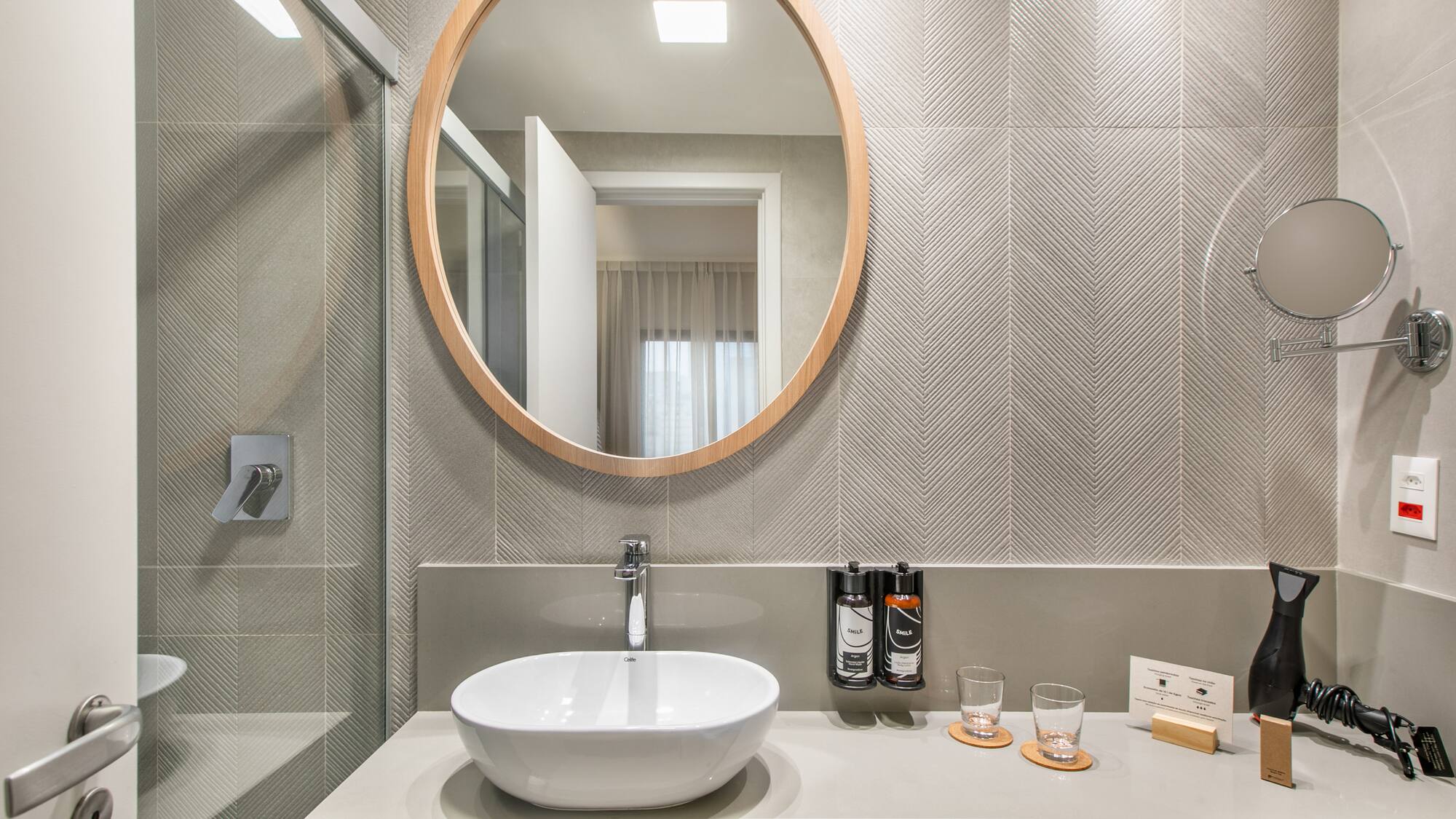 Modern bathroom: round wood mirror, white vessel sink, grey chevron tiles.