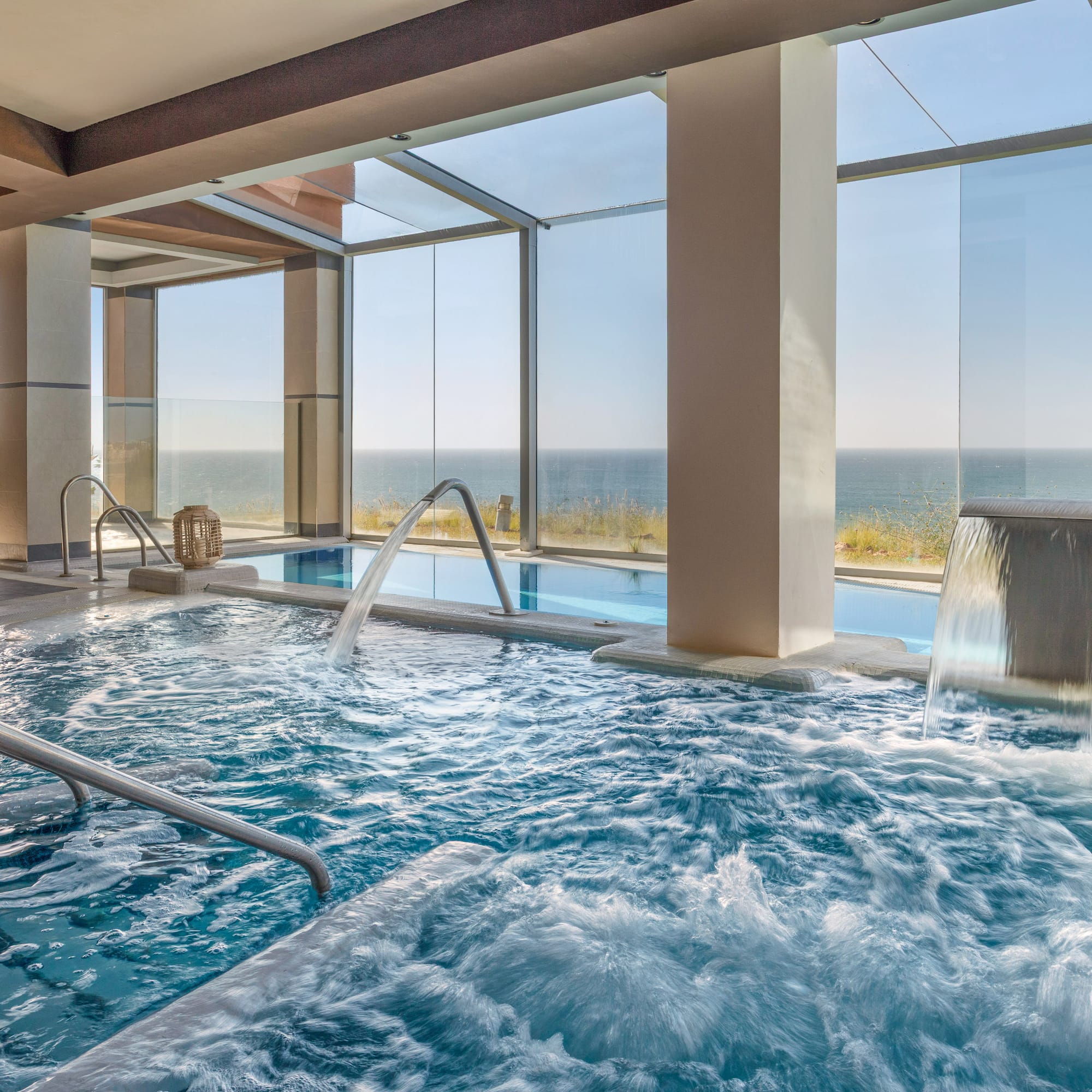 Indoor hydrotherapy spa pool with ocean view, water jets, and waterfall.