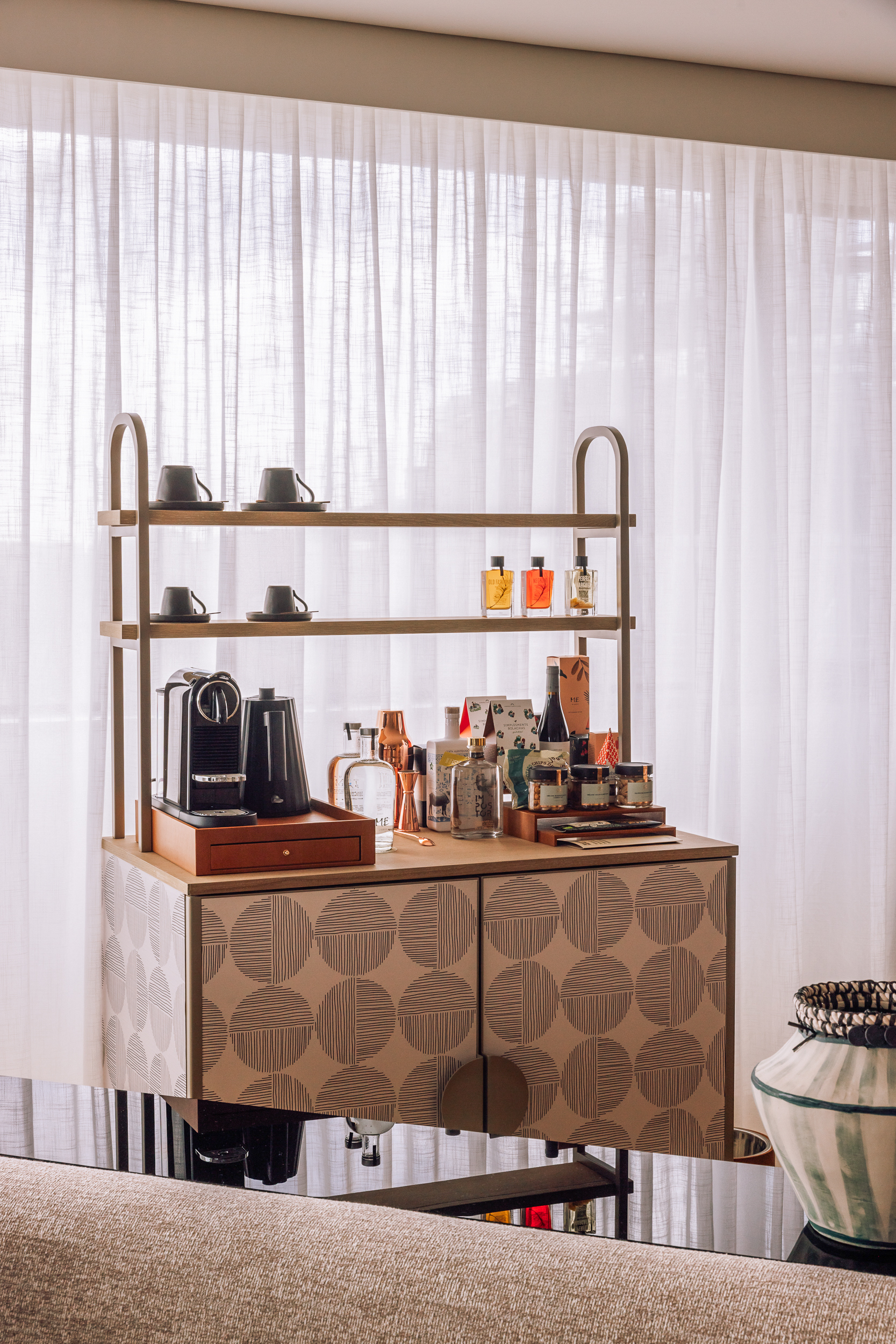 Geometric bar cabinet with coffee maker, liquor, and snacks by sheer curtains.