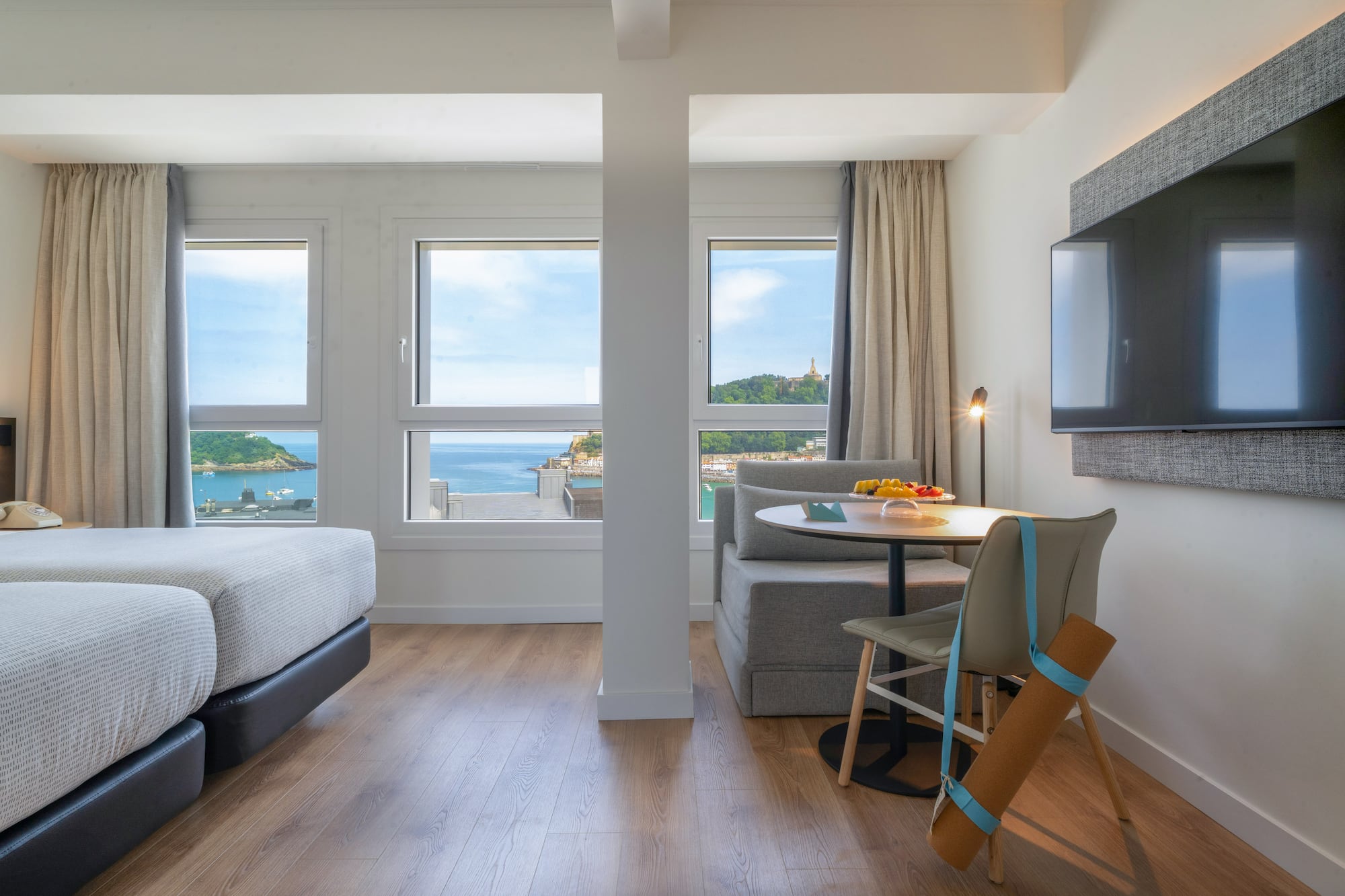 Habitación hotelera con dos camas, vistas a la bahía, sofá y mesa.