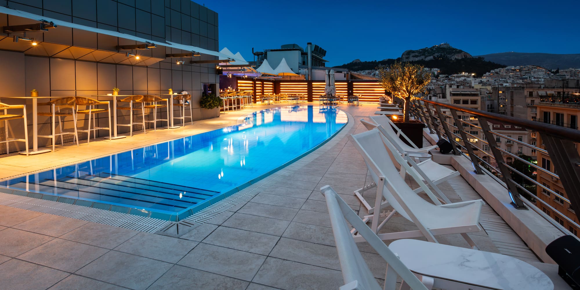 Illuminated Athens rooftop pool at dusk, with city view and bar lounge.