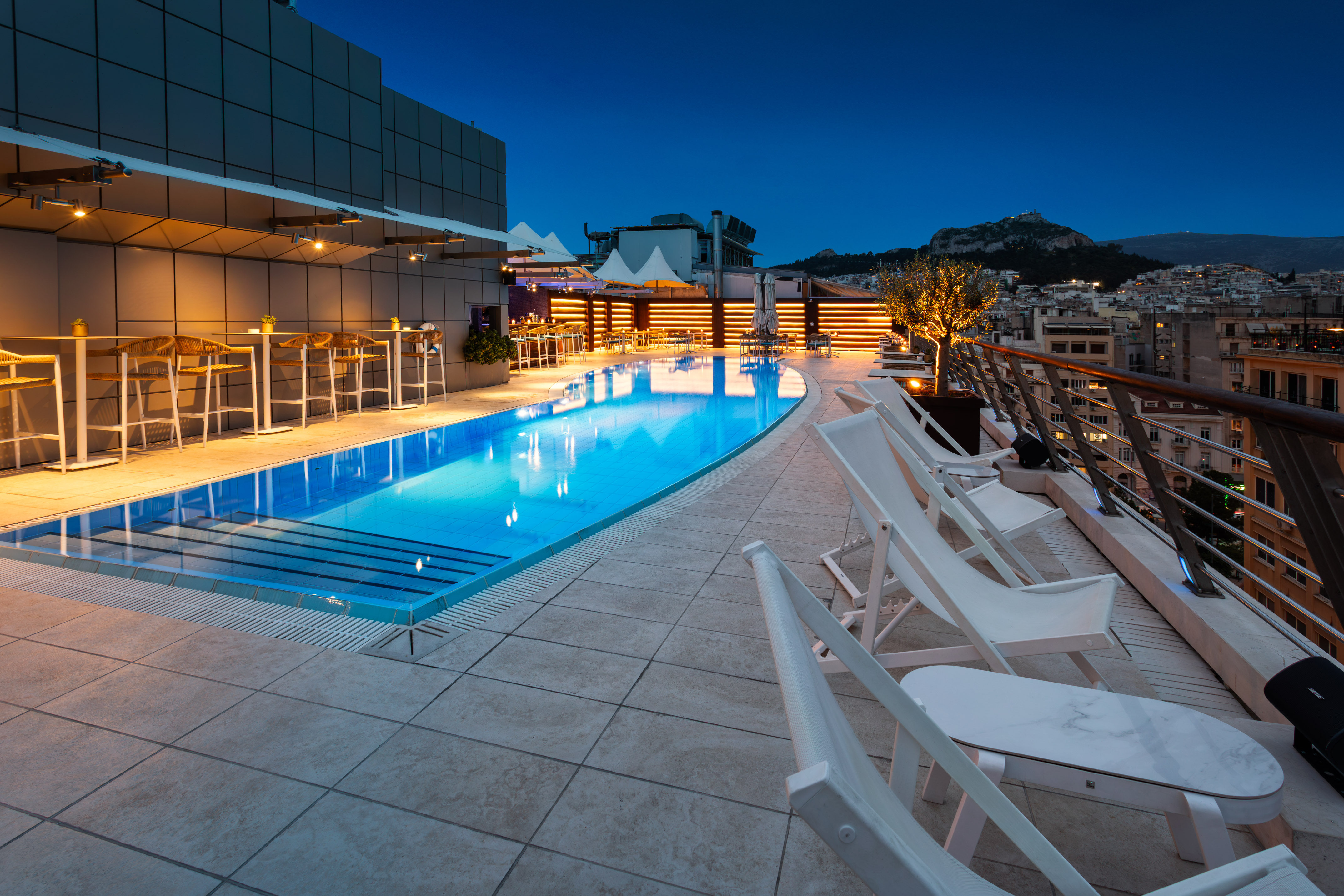 Illuminated Athens rooftop pool at dusk, with city view and bar lounge.