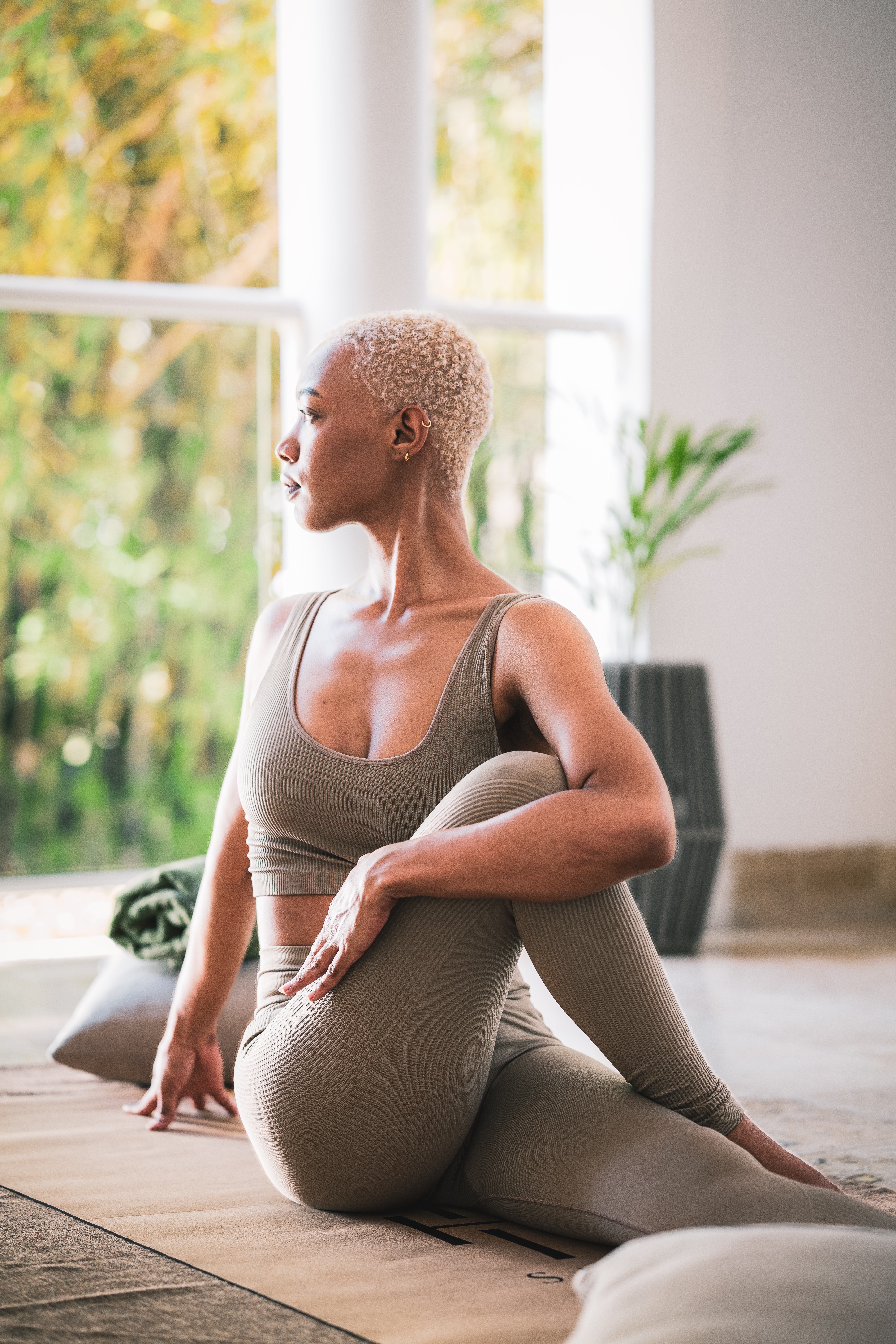 Woman in beige yoga outfit doing seated twist on mat, looking out window.